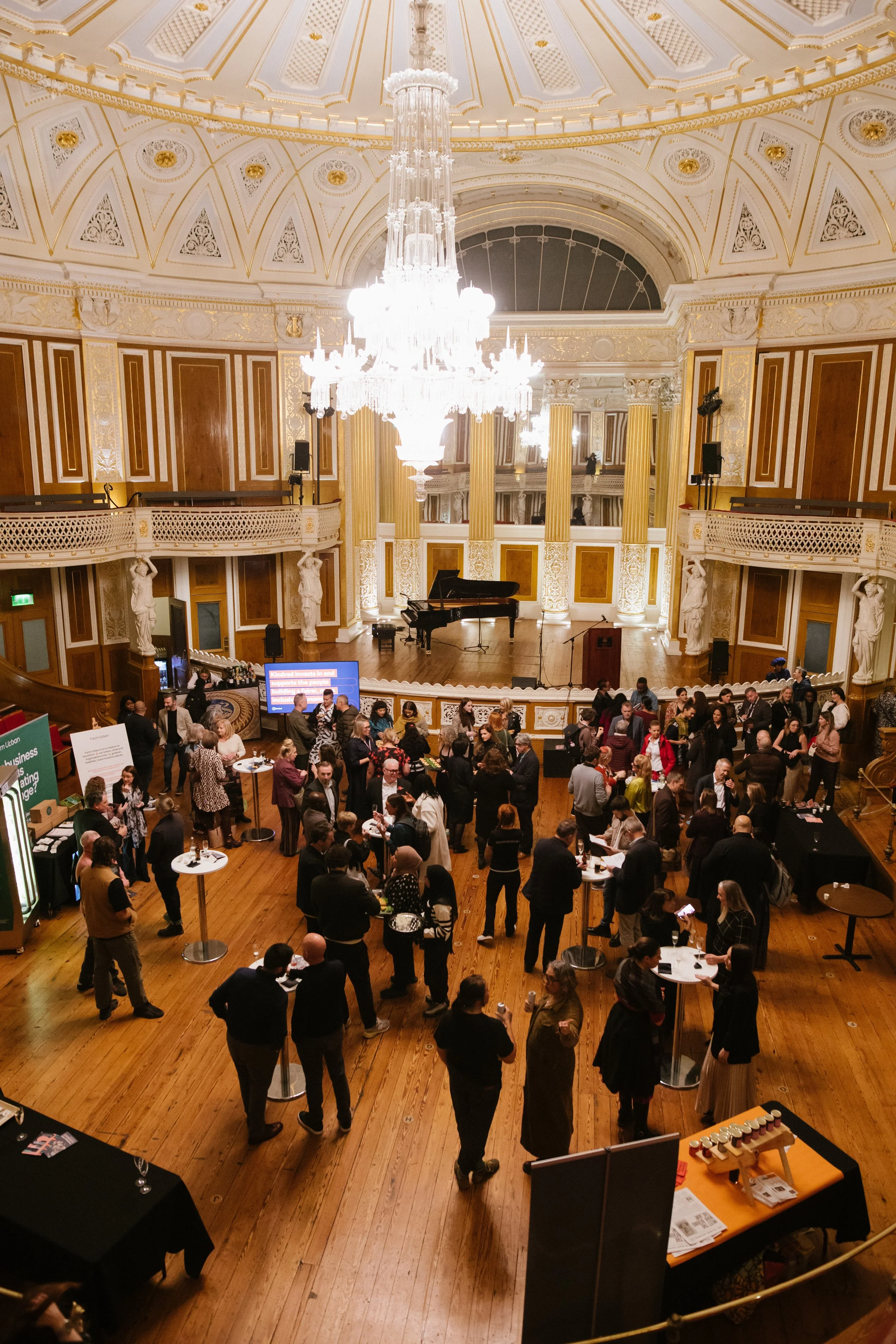 People gathered in a grand, ornate hall with a high, decorated ceiling and a large chandelier, attending a drinks reception with tables, displays, and a stage with a grand piano.
