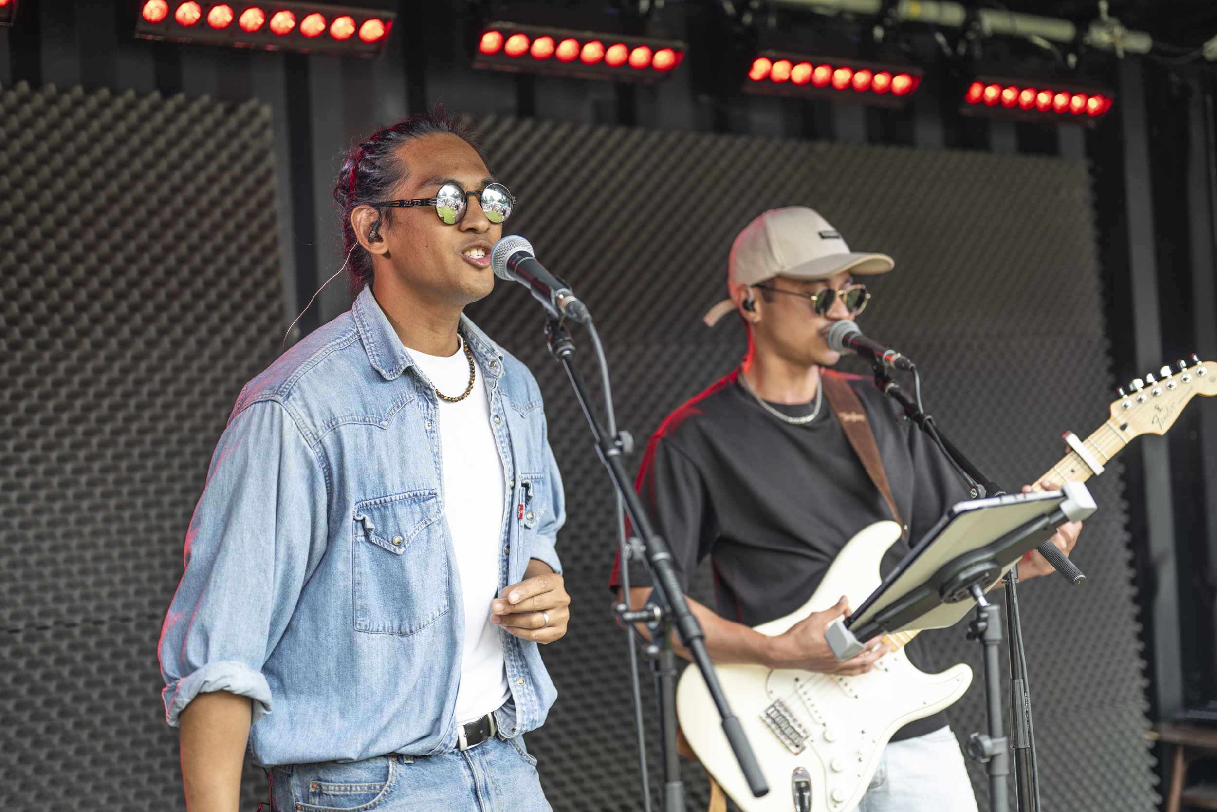 Two male musicians performing on an outdoor stage, one singing into a microphone and the other playing an electric guitar, both wearing sunglasses and casual clothing.
