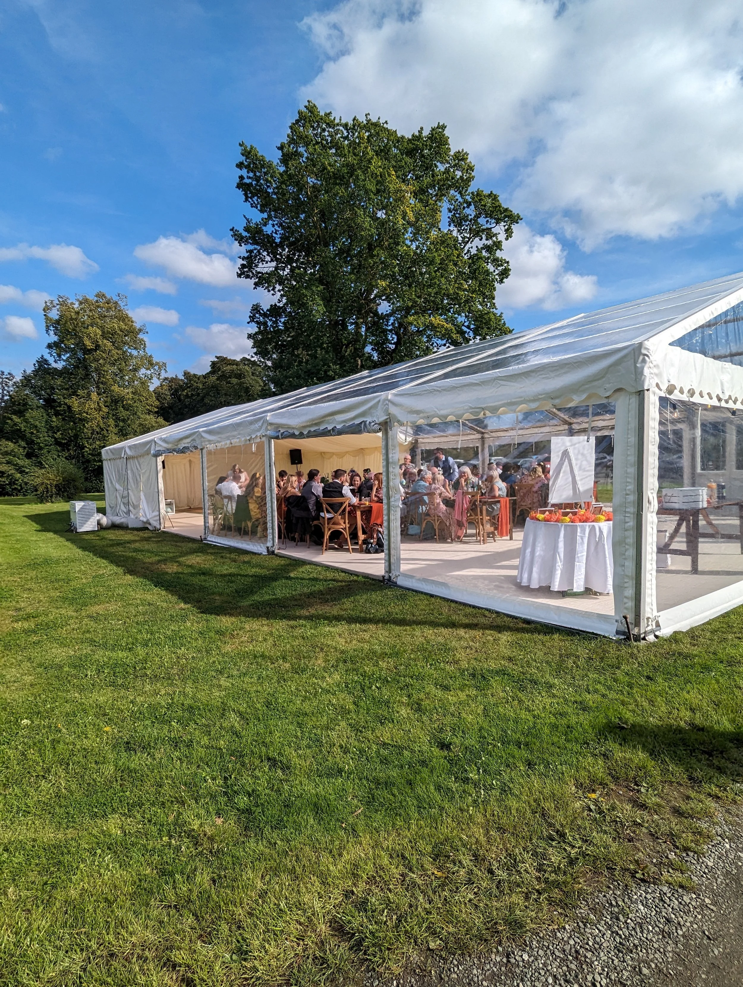 Outdoor event under a white canopy tent with seated guests, decorated tables, and a scenic view of trees and a blue sky with clouds.