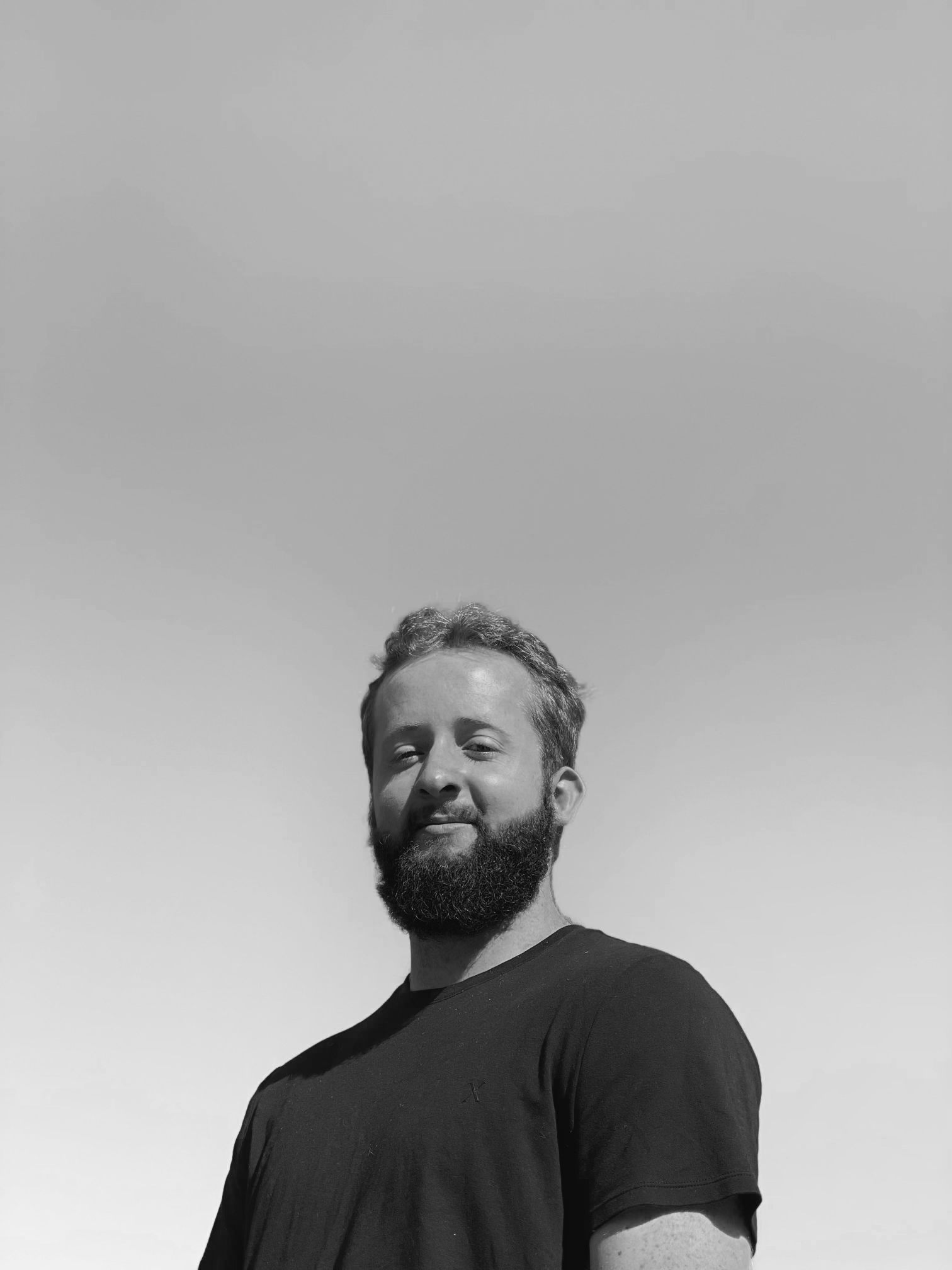 A man with a beard and short hair standing outdoors against a clear sky.