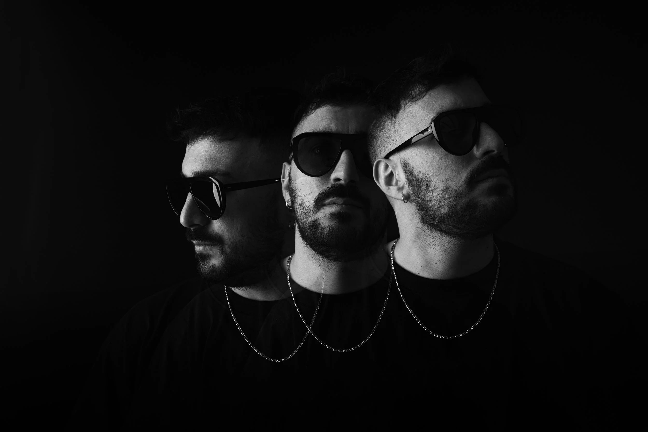 Black and white photo of three men wearing sunglasses and chains, with a dark background, presenting a layered look with each man's face partially overlapping the others.