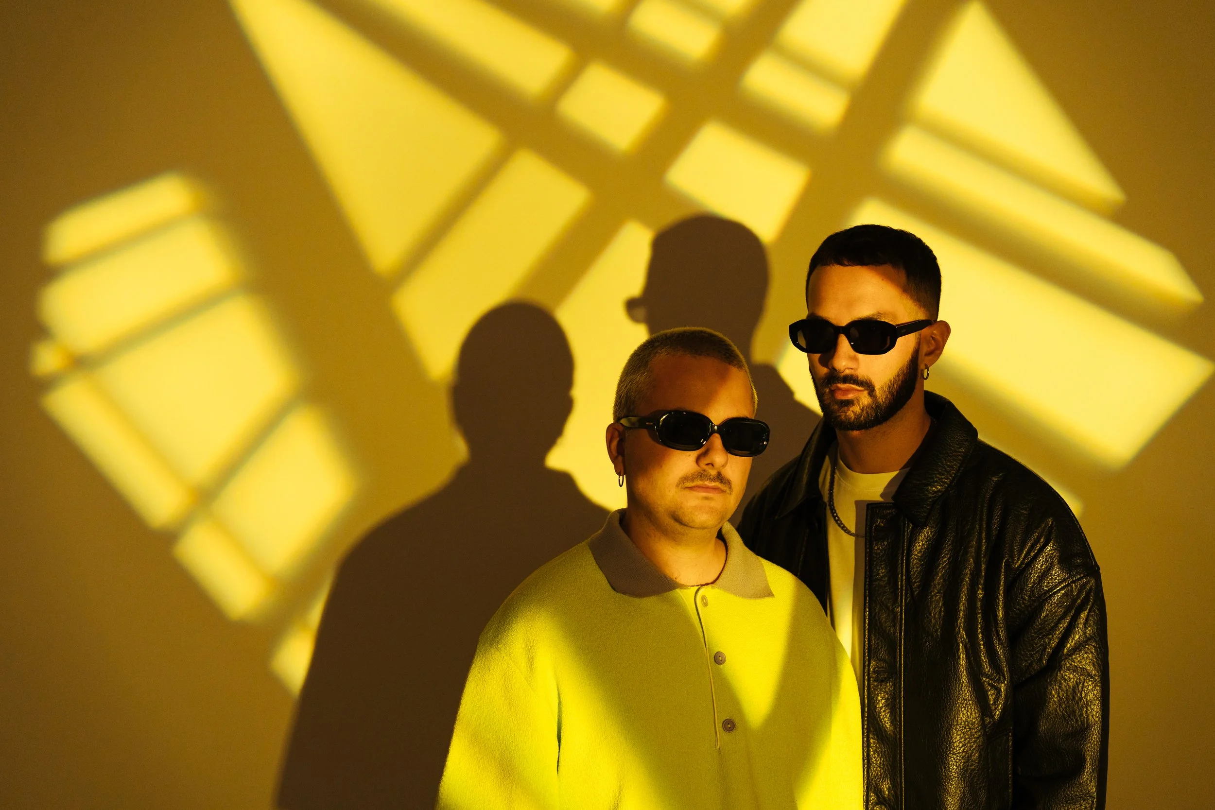 Two men wearing sunglasses stand in front of a yellow wall with projected geometric shadows.