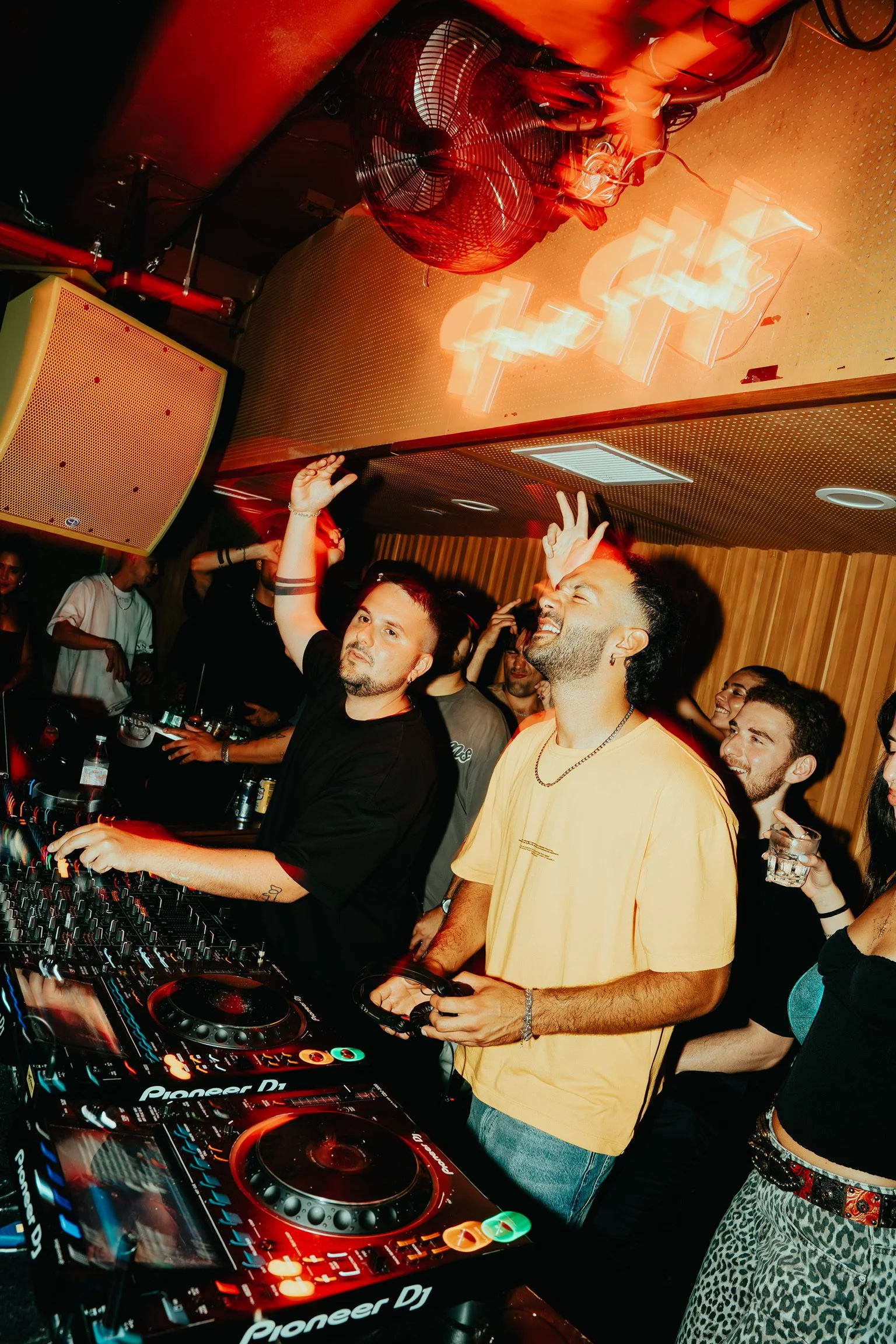 People dancing and enjoying music at a nightclub with DJ equipment, red lighting, and a neon sign on the wall.