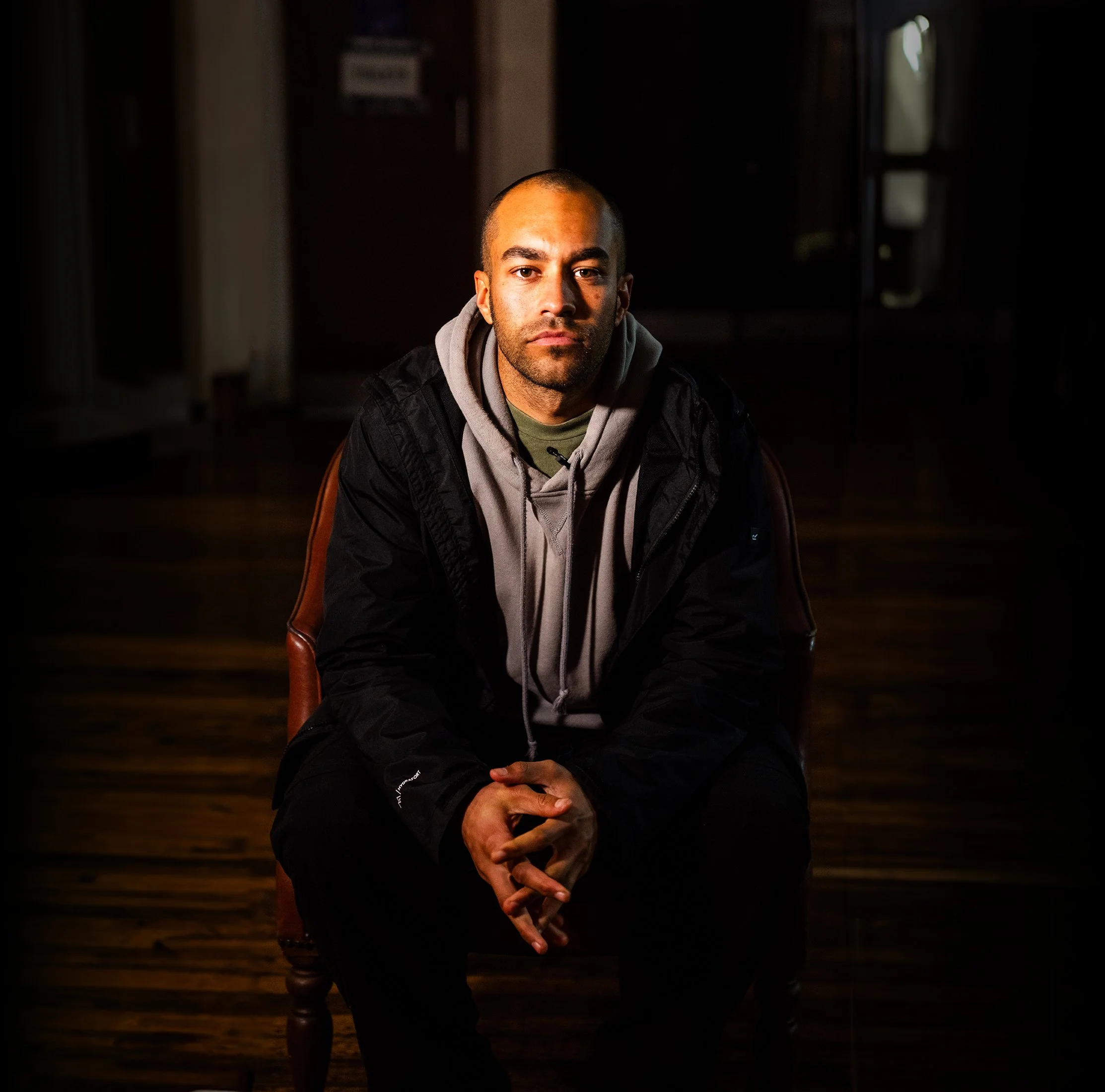 A young man sitting on a chair in a dimly lit room, wearing a black jacket, gray hoodie, and green shirt, with a serious expression.