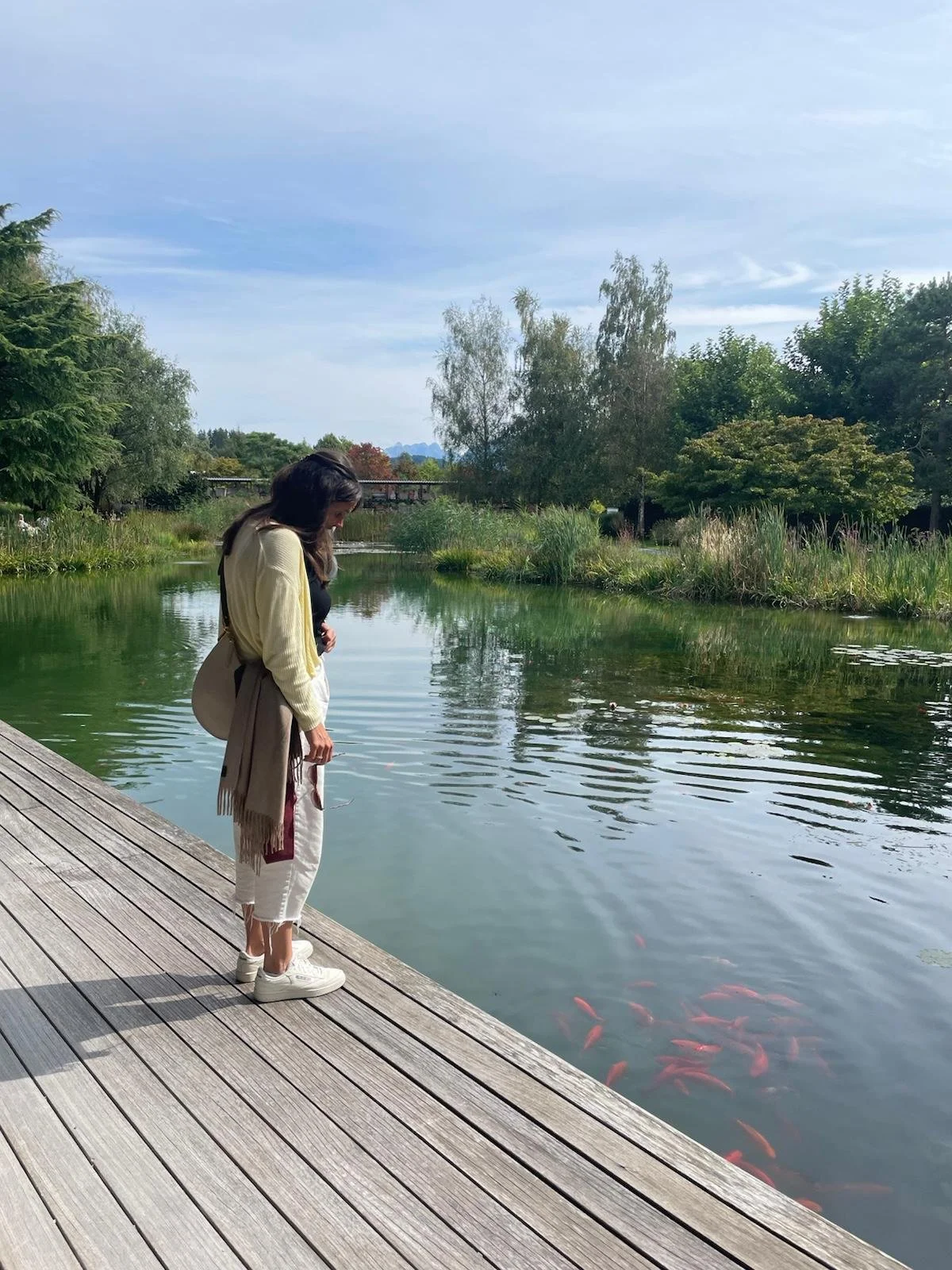 A woman in casual clothing standing on a wooden dock, looking into a pond with colorful fish, surrounded by trees and bushes under a partly cloudy sky.