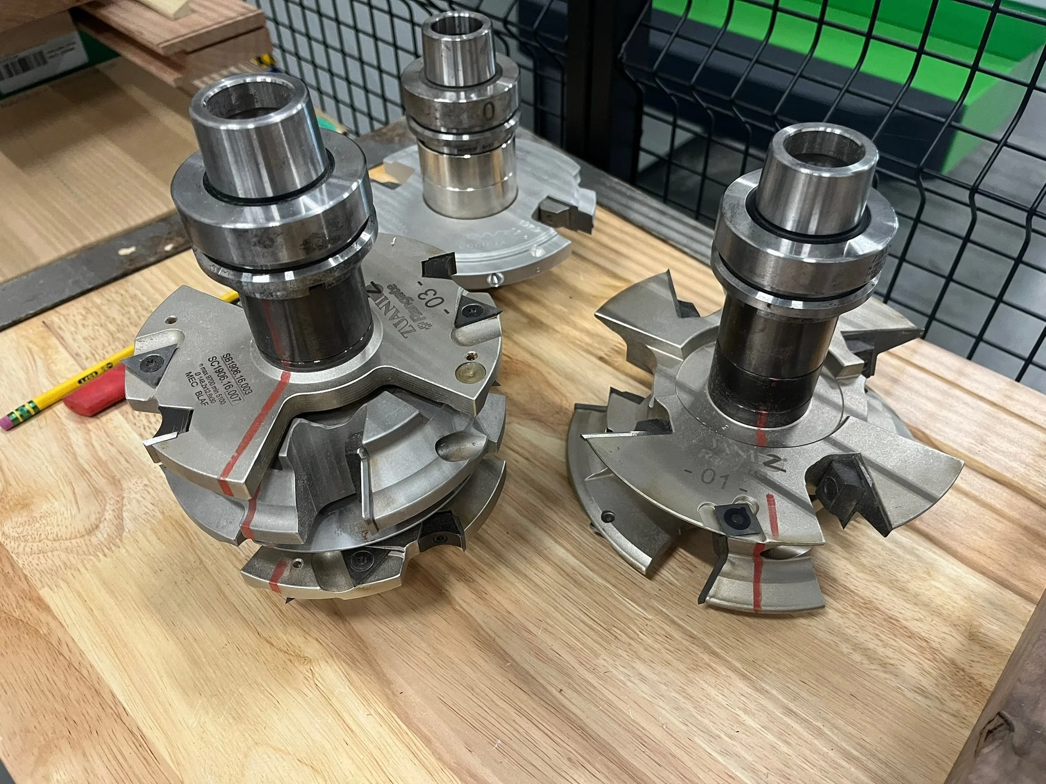 Close-up of three metal industrial machine parts on a wooden surface. They are cylindrical with multiple cutouts and markings, and are used for manufacturing or machining processes with a CNC router.