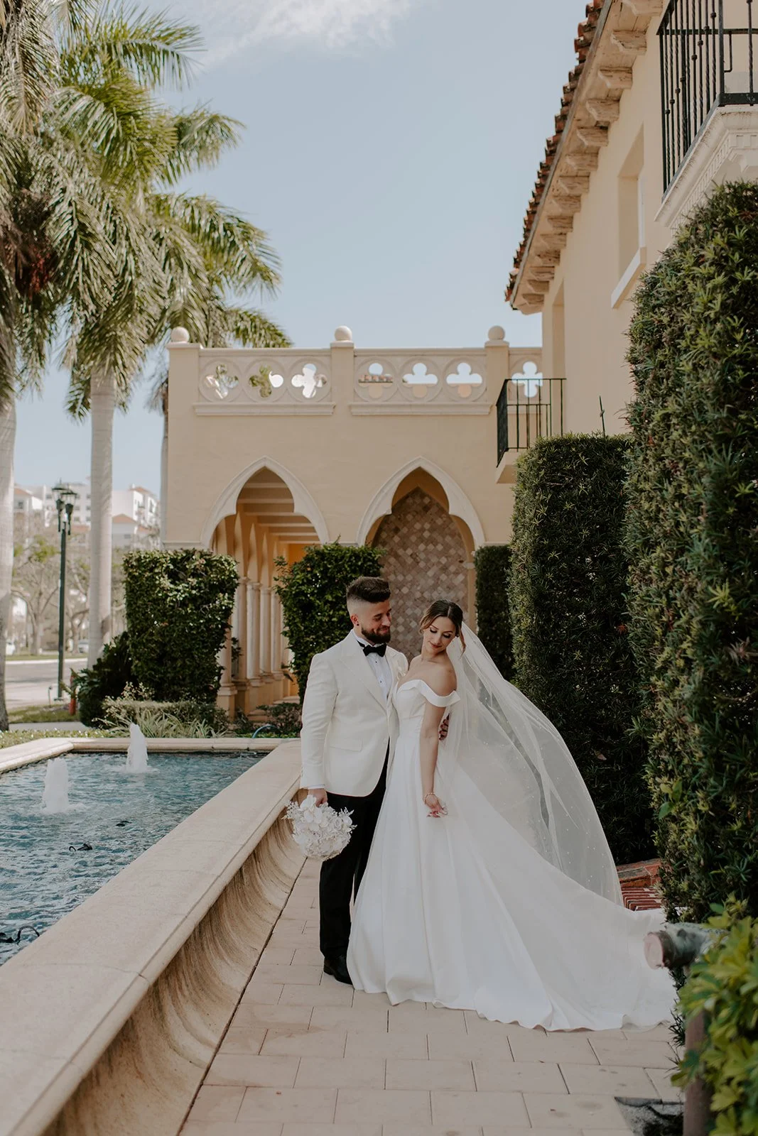 A bride and groom in wedding attire standing beside a fountain in a lush garden, with a beige building featuring arches and balconies in the background, under a clear sky.