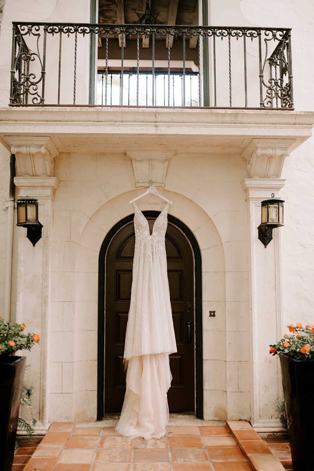 A wedding dress hanging on a hanger in front of a dark wooden door, with a stone archway above it, flanked by two wall-mounted lanterns, with potted plants with orange flowers on either side.