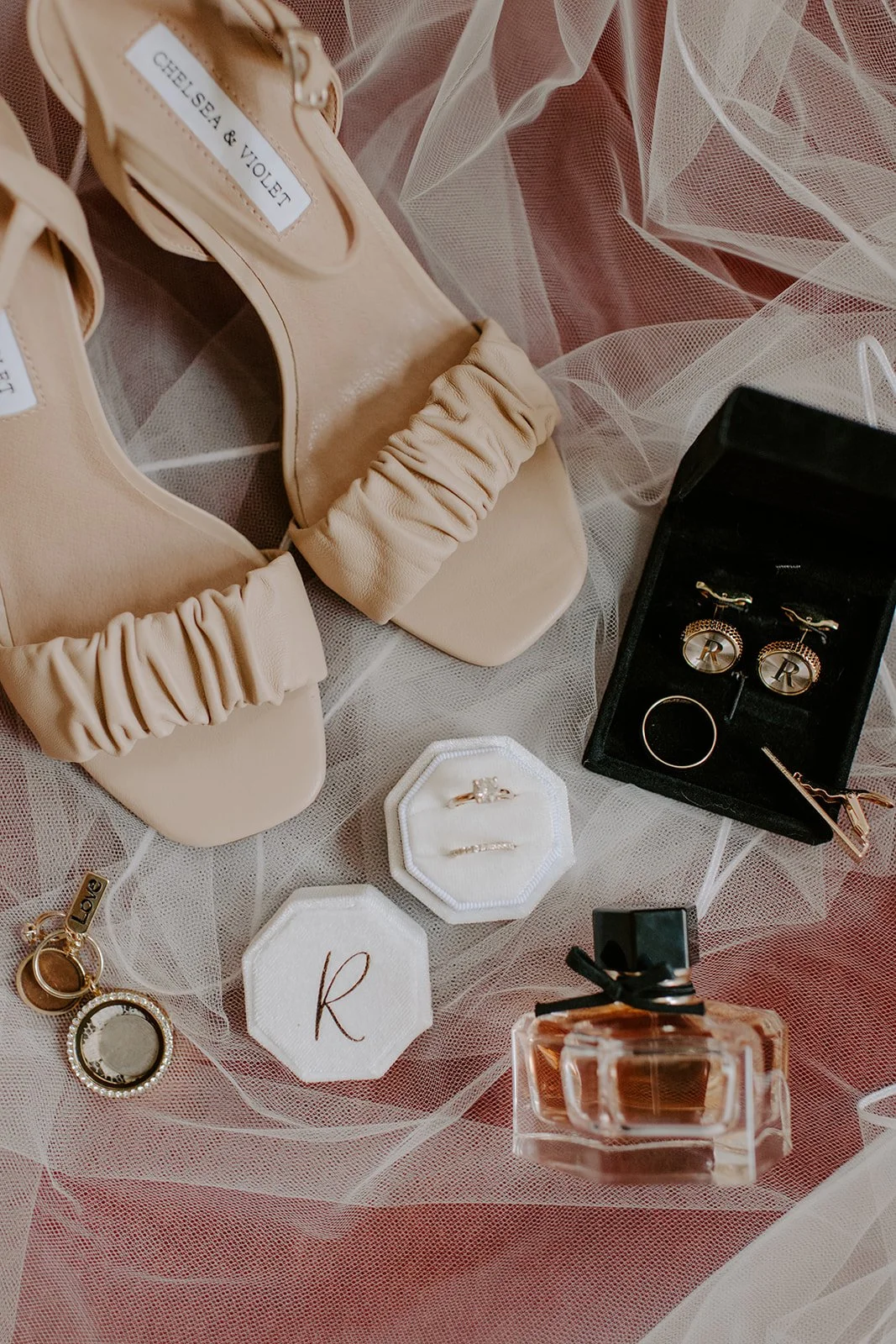 Beige David's Bridal wedding shoes, jewelry box with rings, earrings in a black jewelry box, perfume bottle, and wedding ring on sheer pink fabric.