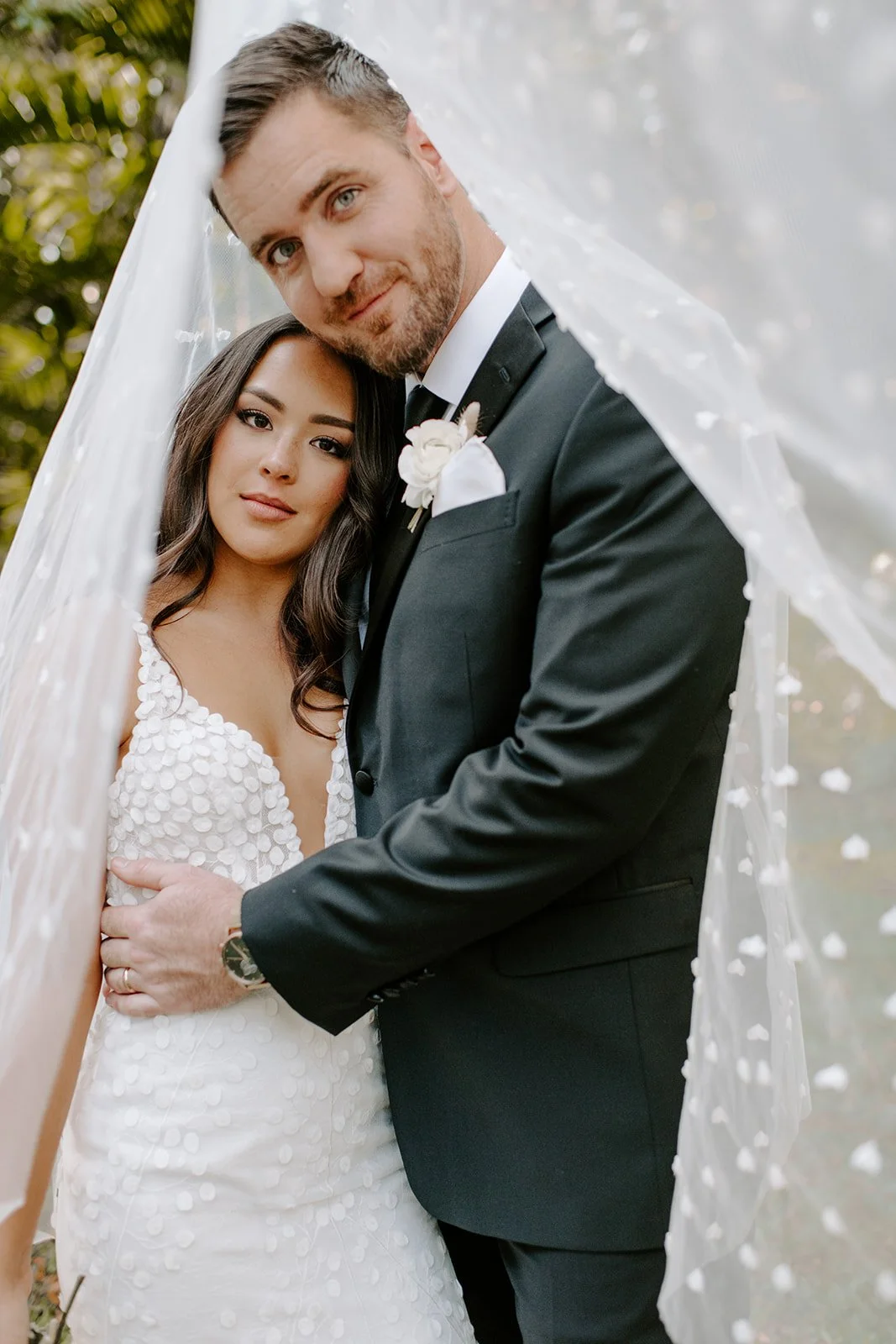 A bride and groom pose under a white veil, with the bride in a white dress and the groom in a black tuxedo, outdoors with greenery in the background.
