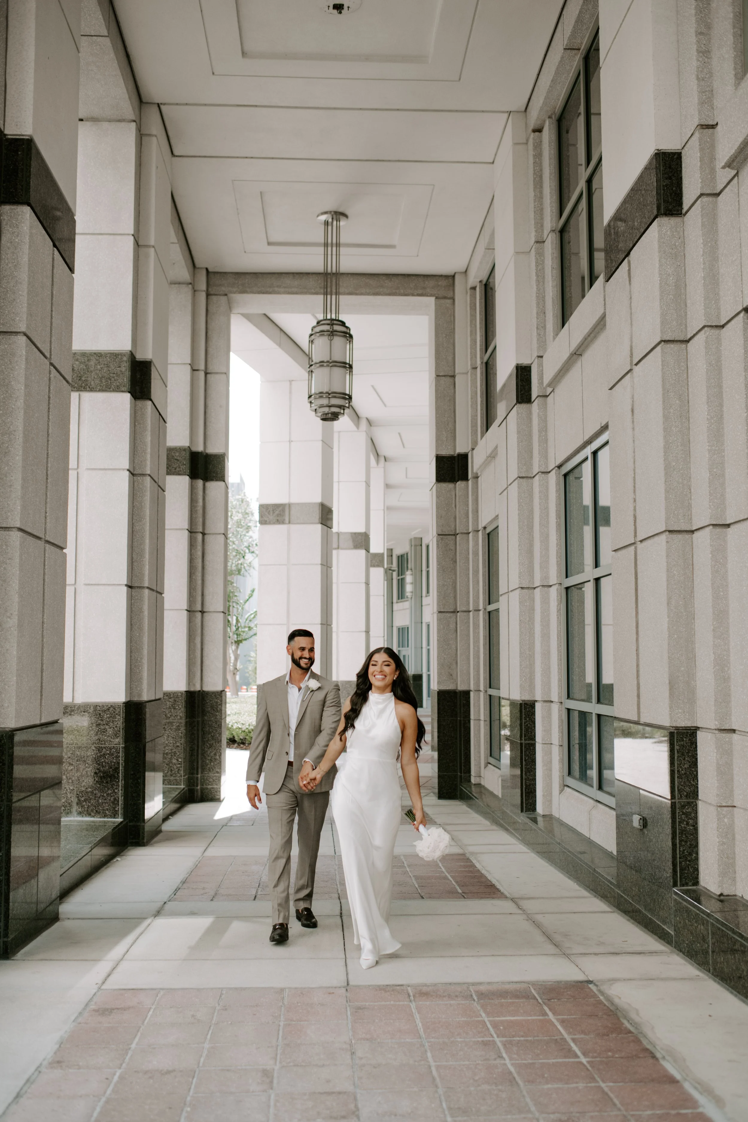 A happy bride and groom walking hand in hand in a modern outdoor corridor, smiling and holding a bouquet of flowers.