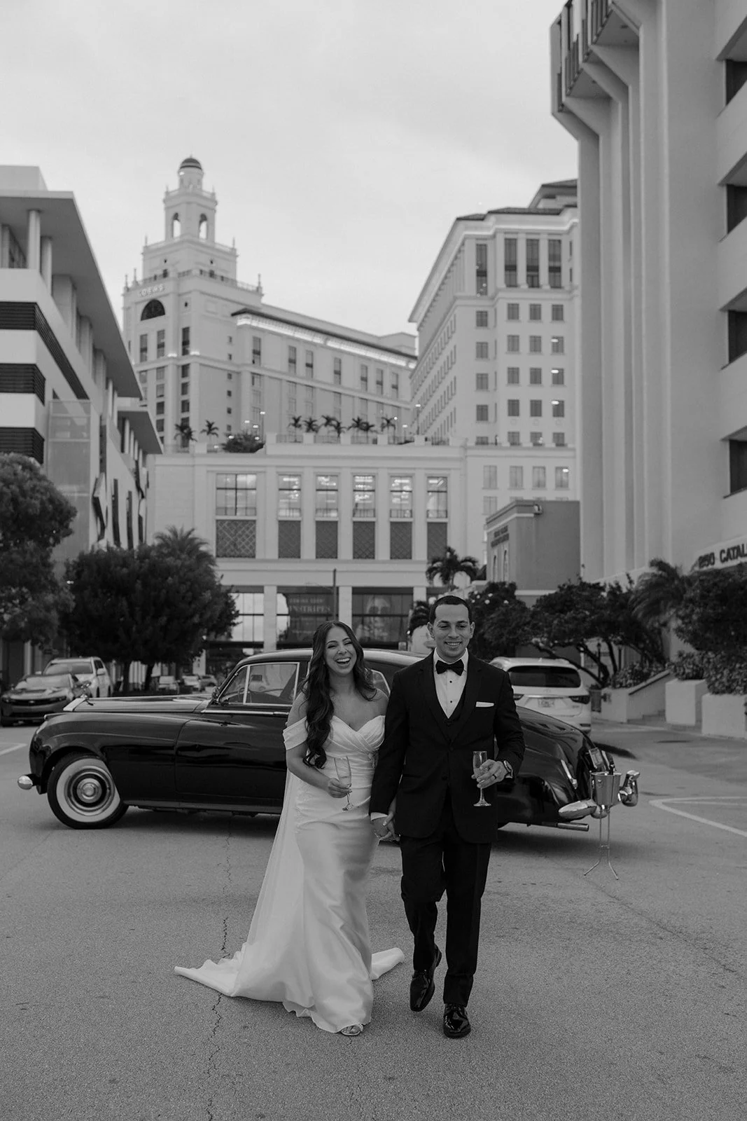 A newlywed couple in formal attire walking hand in hand on a city street, with a vintage black car in the background, tall buildings, and trees.