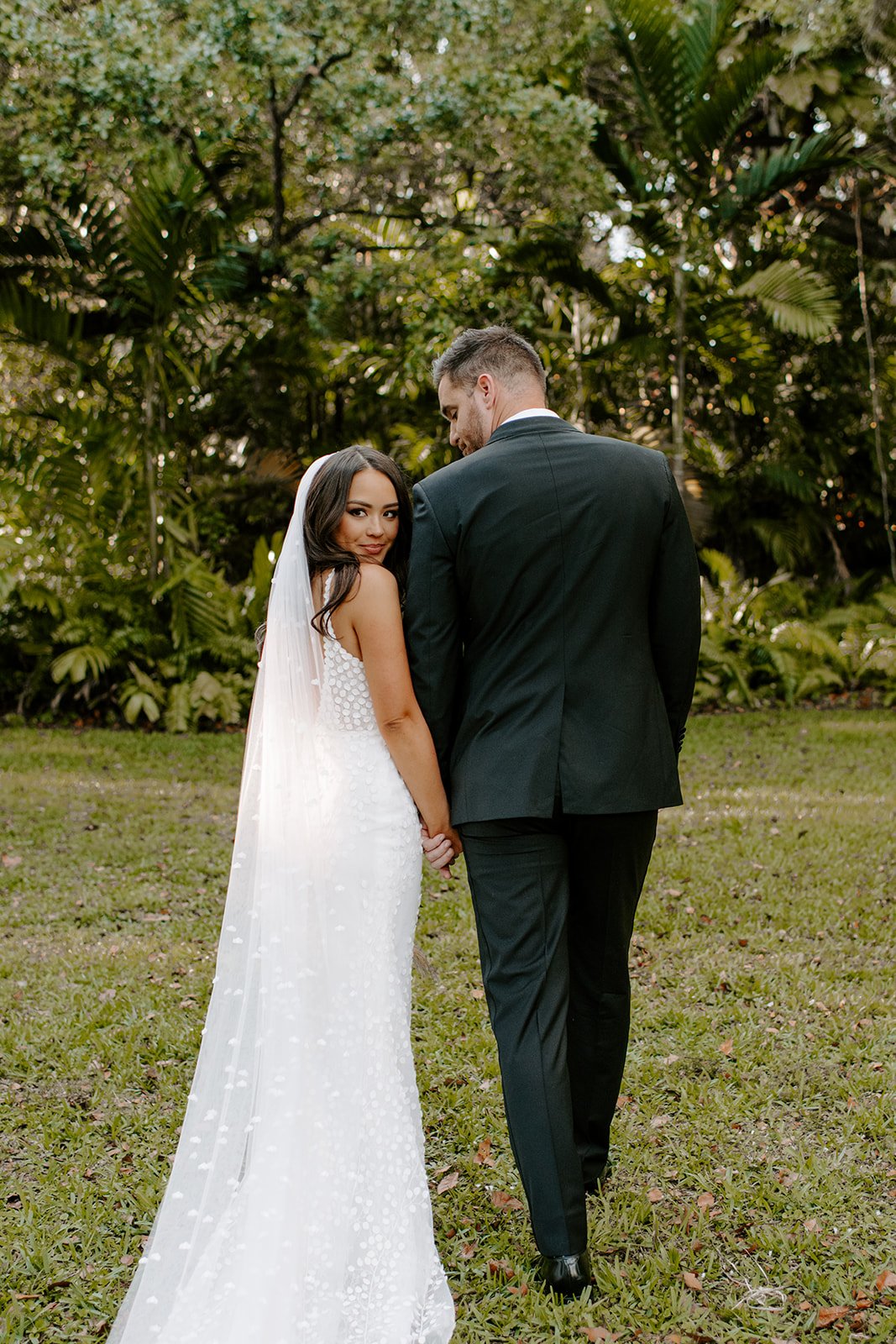 A bride and groom holding hands outdoors in a green garden, with the bride looking over her shoulder at the camera.
