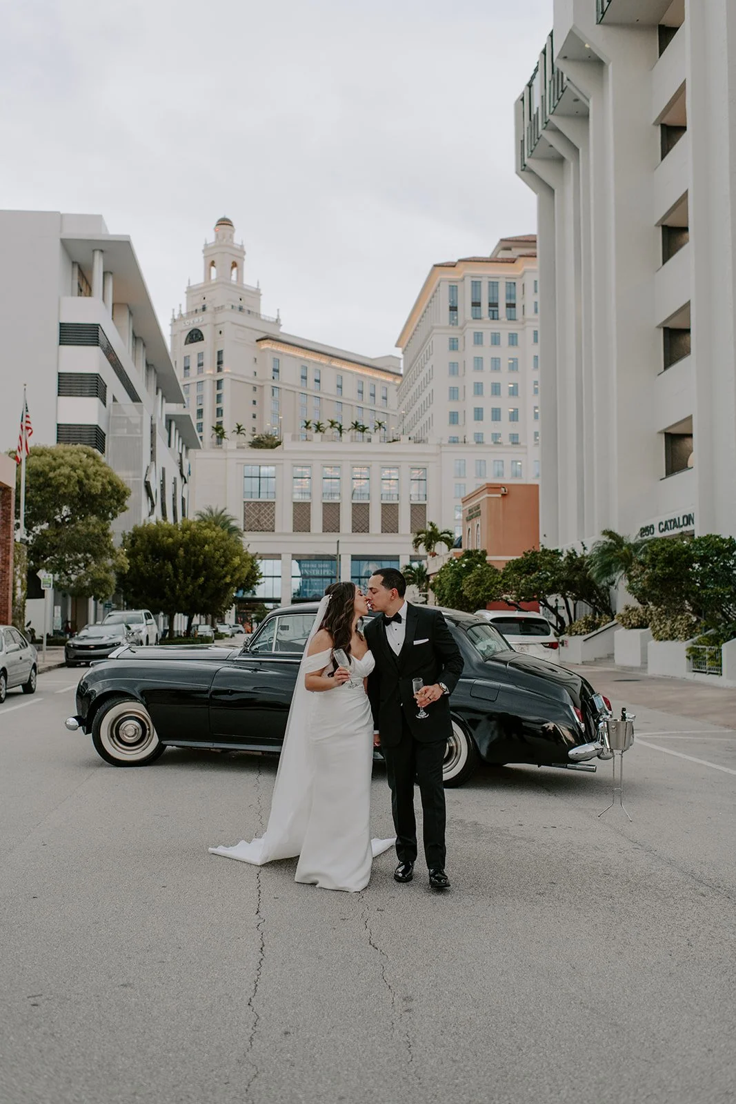 A newlywed couple in wedding attire sharing a kiss in front of a vintage black car on a city street, with tall white buildings and palm trees in the background.