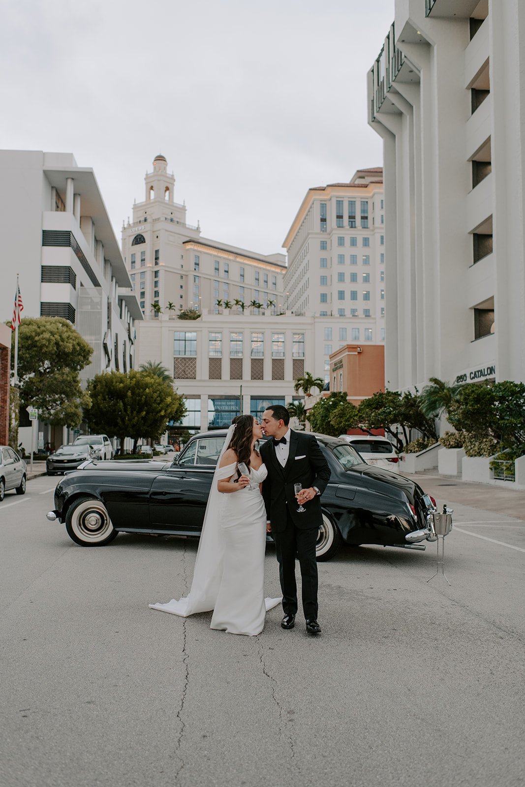 A newly married bride and groom kissing in front of a vintage black car on a city street, with tall white buildings and palm trees in the background.