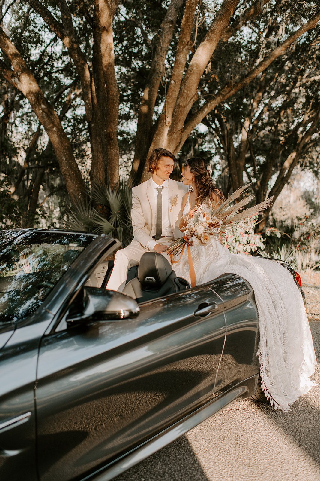 A newlywed couple sitting on the back of a black convertible car outdoors, smiling at each other, with large trees and sunlight filtering through the leaves in the background. The bride is holding a bouquet of flowers.