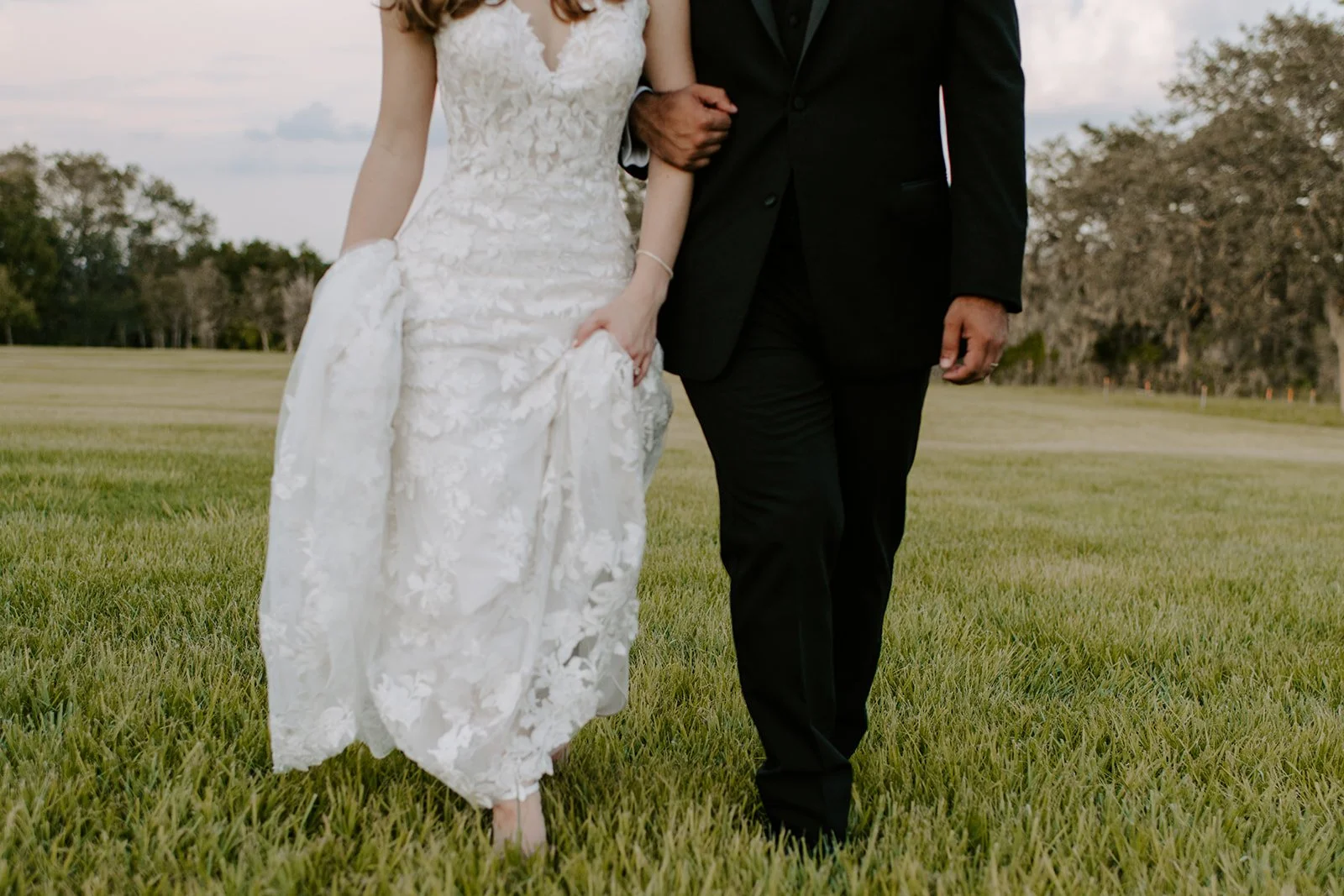 A bride in a white lace wedding gown holding up her dress, walking arm-in-arm with a groom in a black suit, on a grassy field with trees in the background.
