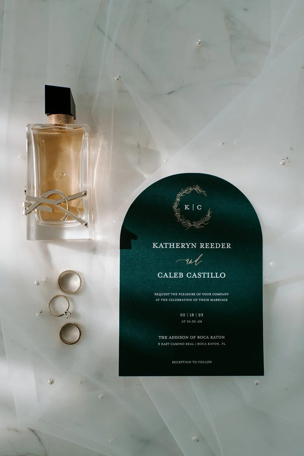 Wedding invitation card with names Katheryn Reeder and Caleb Castillo, wedding date 02/18/23 at 10:30 AM, location at The Addison of Boca Raton, surrounded by rings, perfume bottle, and decorative tulle fabric on a white surface.