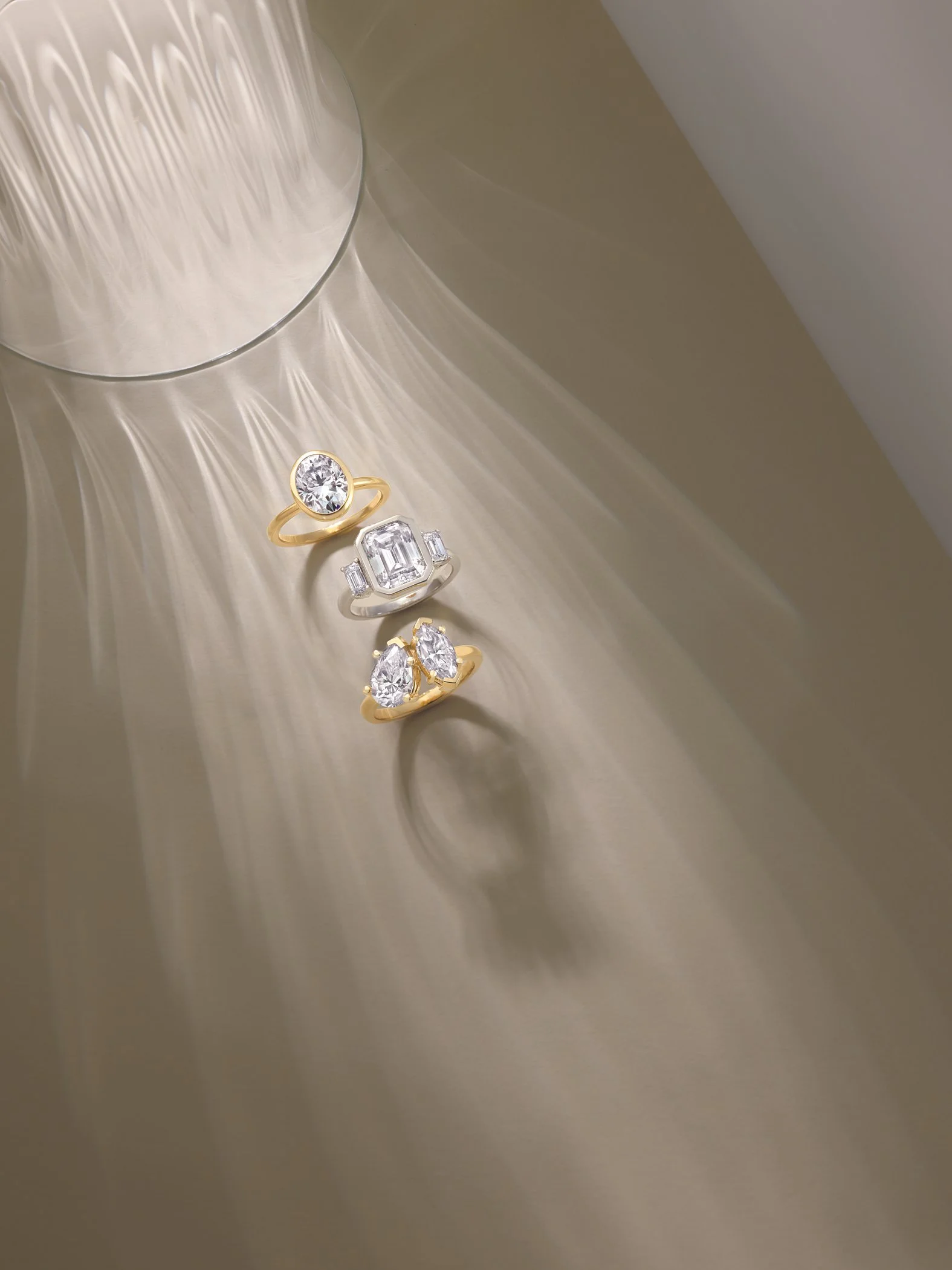 Three diamond rings with gold and silver bands are placed on a gold-colored surface, with a reflective bowl nearby casting reflections and light patterns.