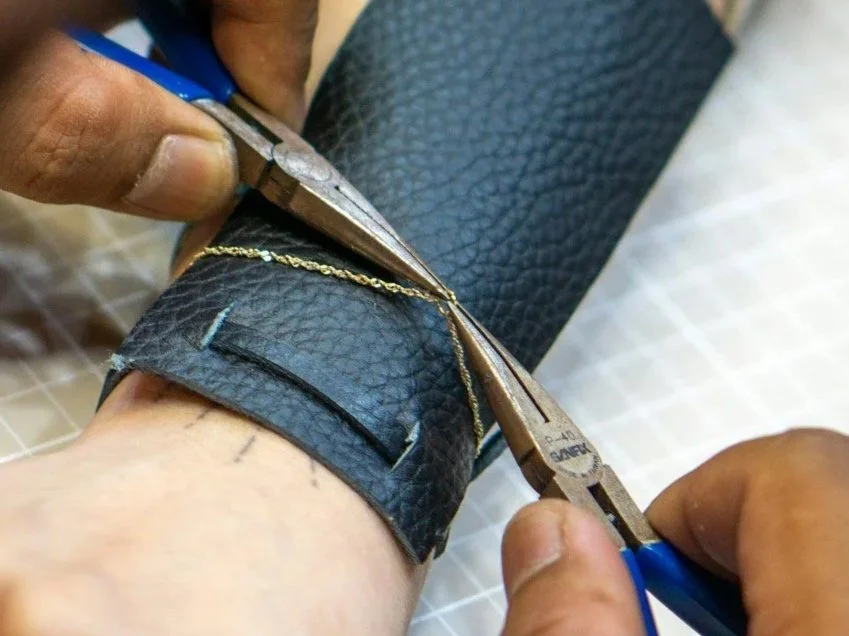 Close-up of a person doing permanent jewelry using pliers and scissors to cut a gold chain on a leather wristband.