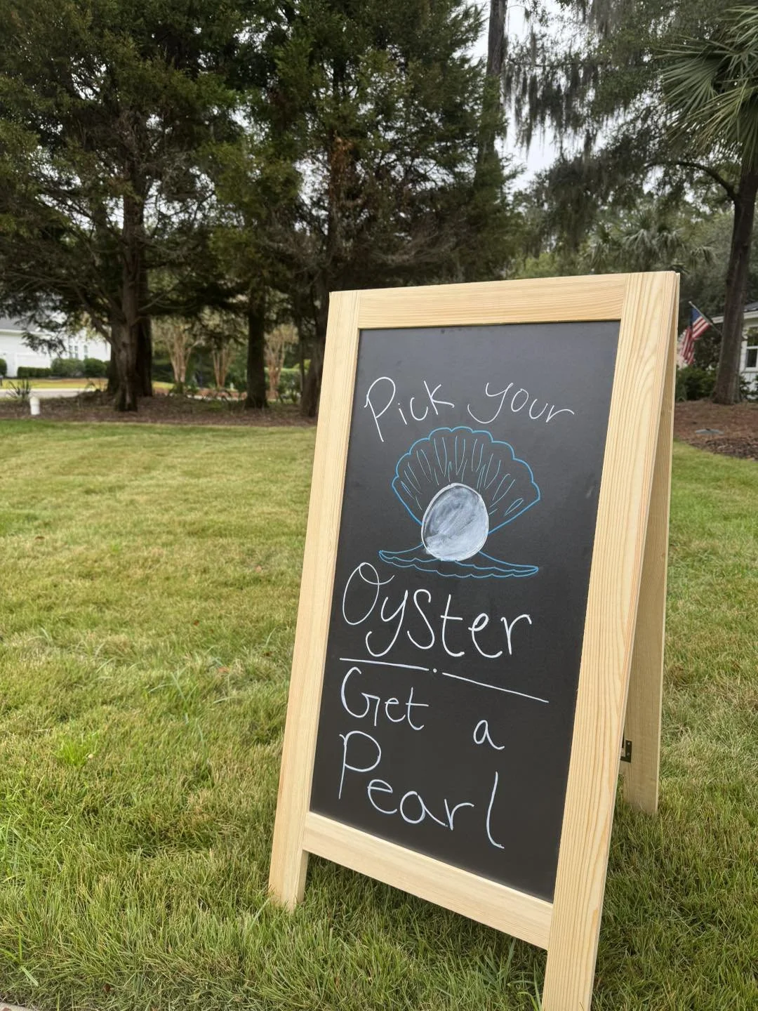 A chalkboard sign outdoors with a drawing of an oyster shell and the song lyrics 'Pick your Oyster, Get a Pearl' written on it, placed on grass with trees and a house in the background.