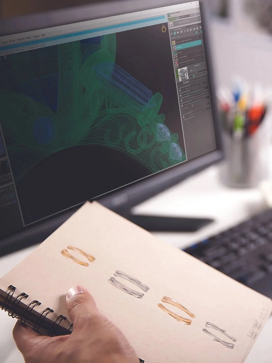 Person holding a spiral notebook with various colored sketches of rings in front of a computer monitor displaying 3D design software with wireframe models.