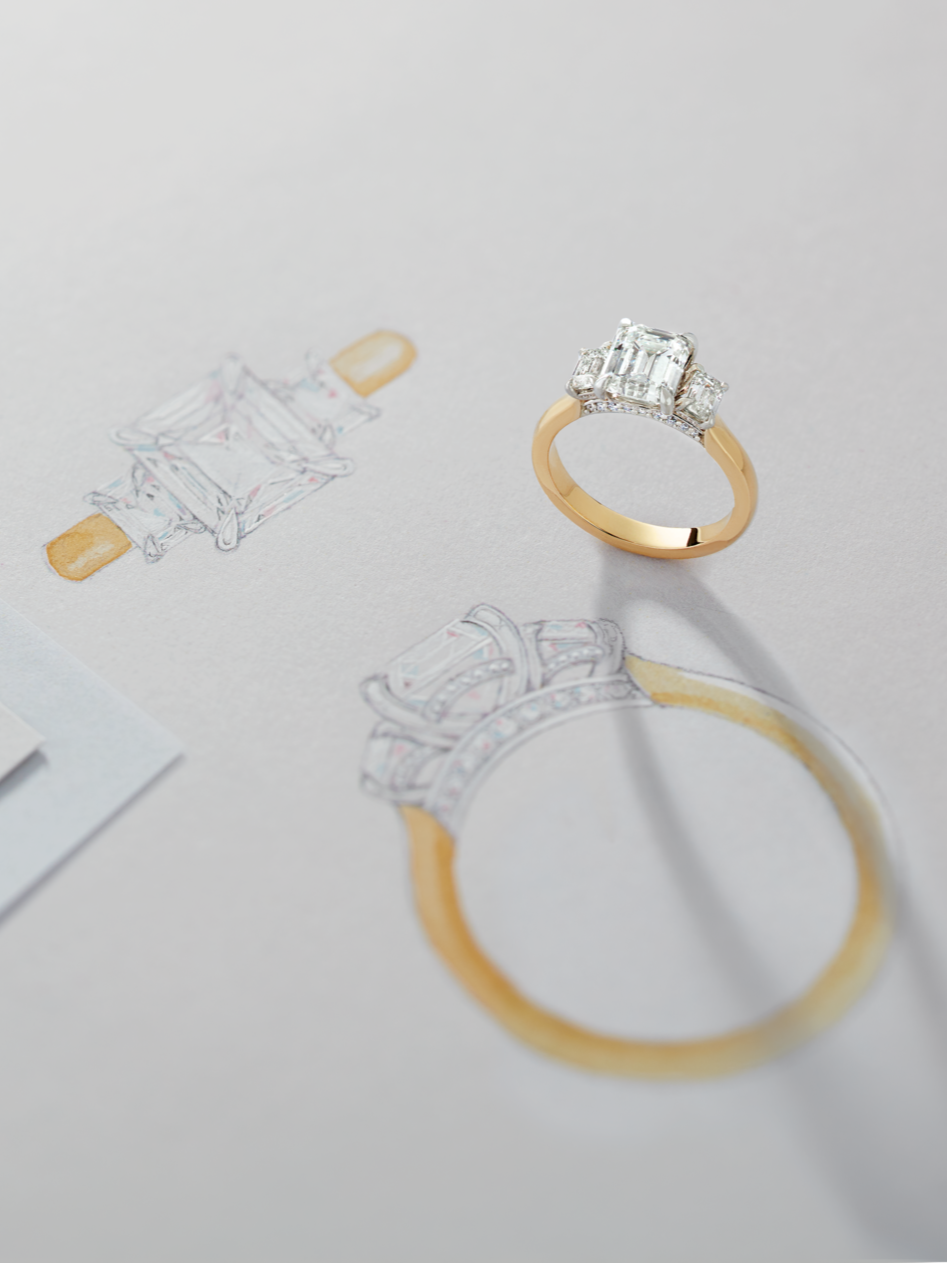 A yellow gold ring with a three-stone emerald engagement ring on white paper with design sketches of the yellow gold ring.