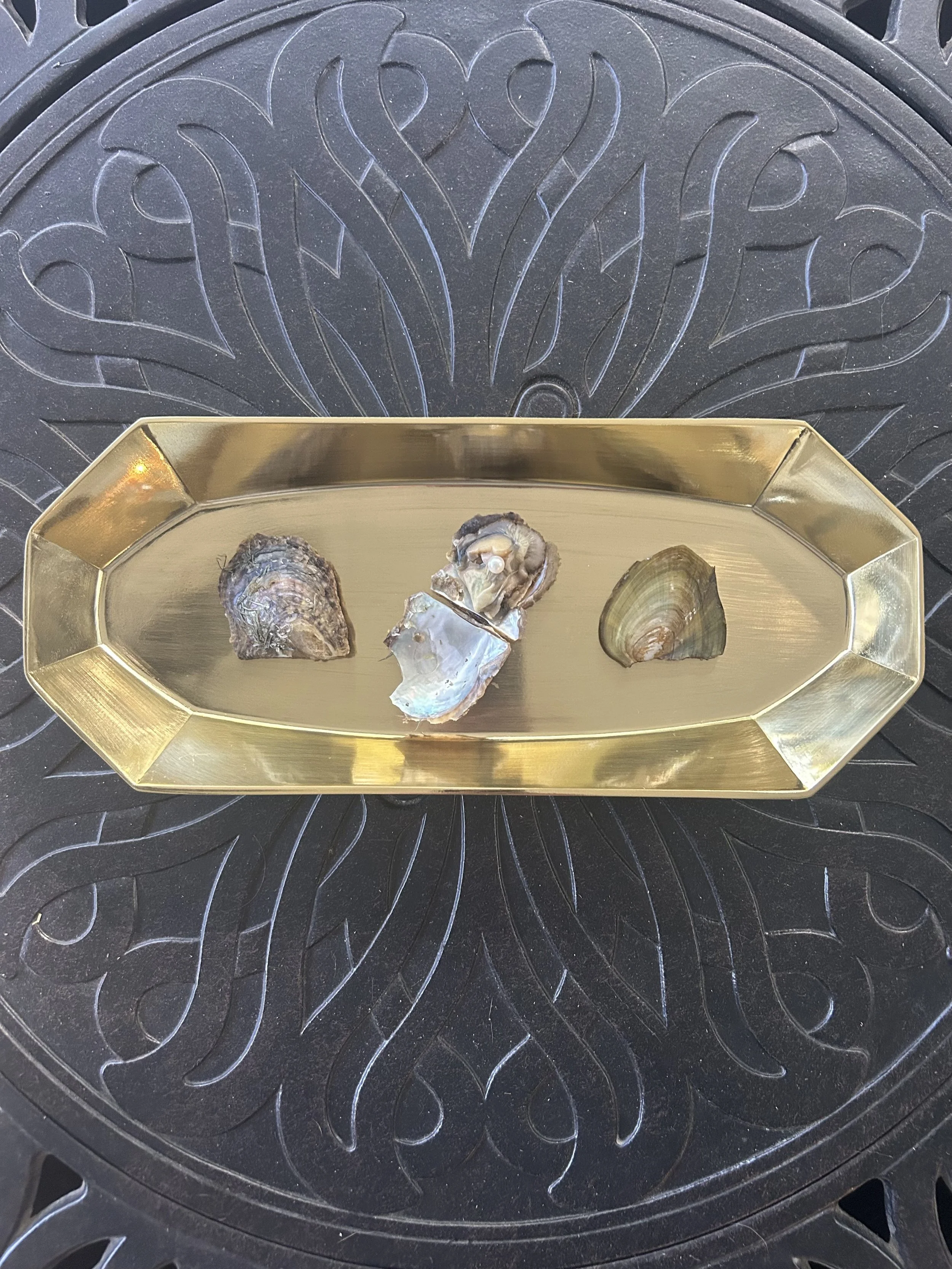 Three oyster shells with a pearl on a gold rectangular tray, sitting on a black patterned surface.