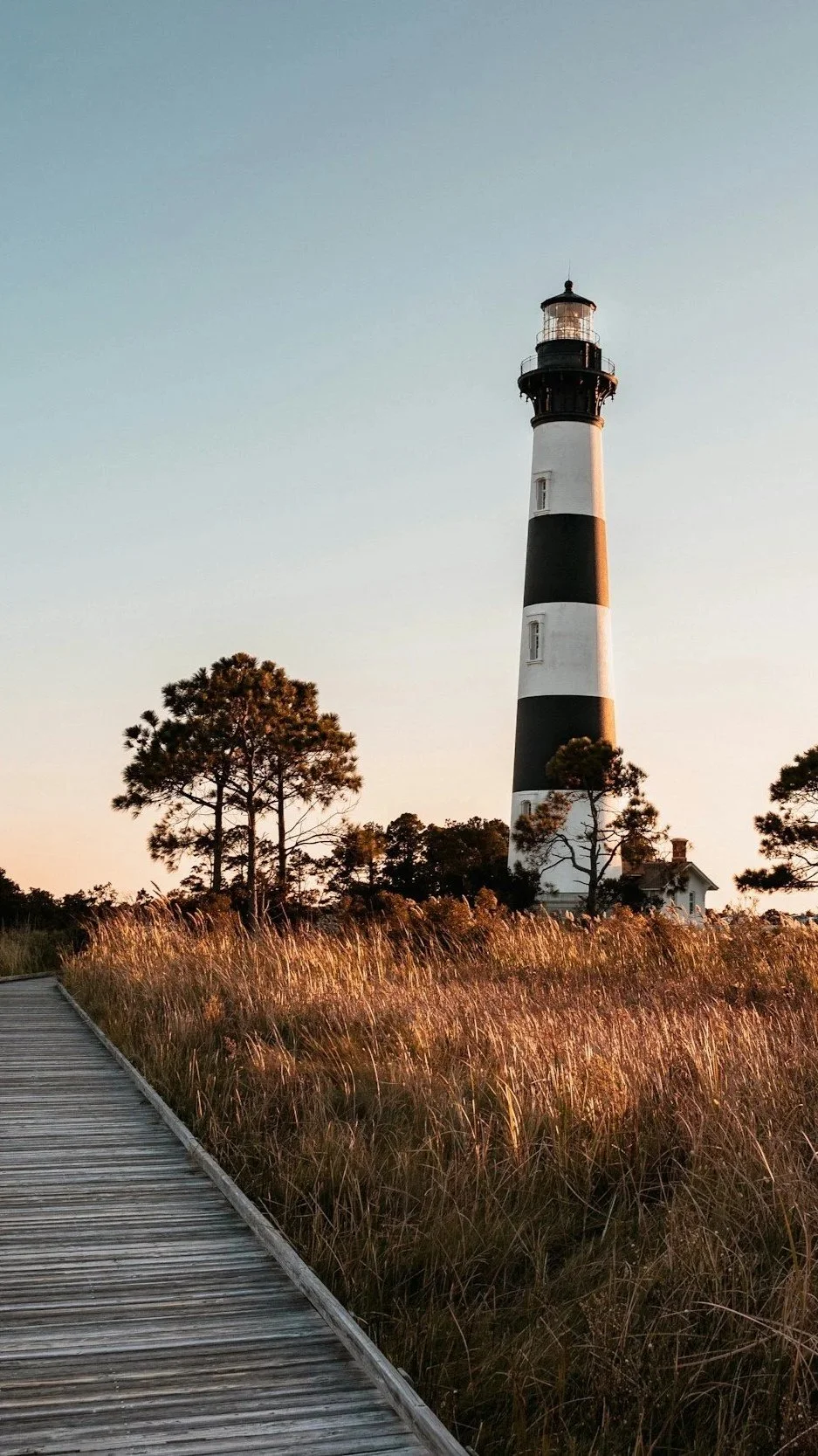 A lighthouse with black and white stripes standing on a grassy area with trees, a wooden walkway in the foreground, and a clear sky.