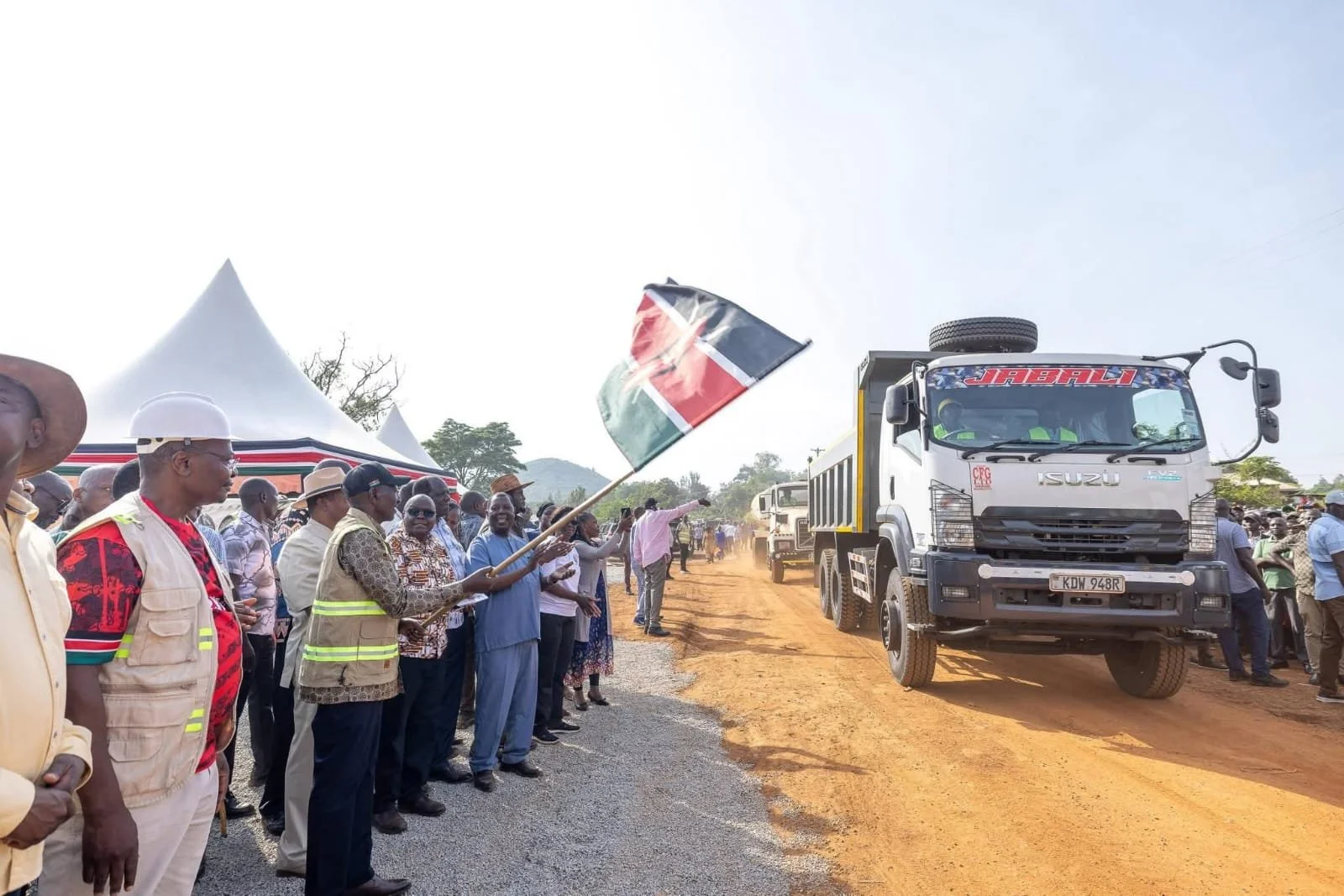 Crowds stand along the roadside and wave the Kenya flag as large trucks carry transport on the dirt roads.