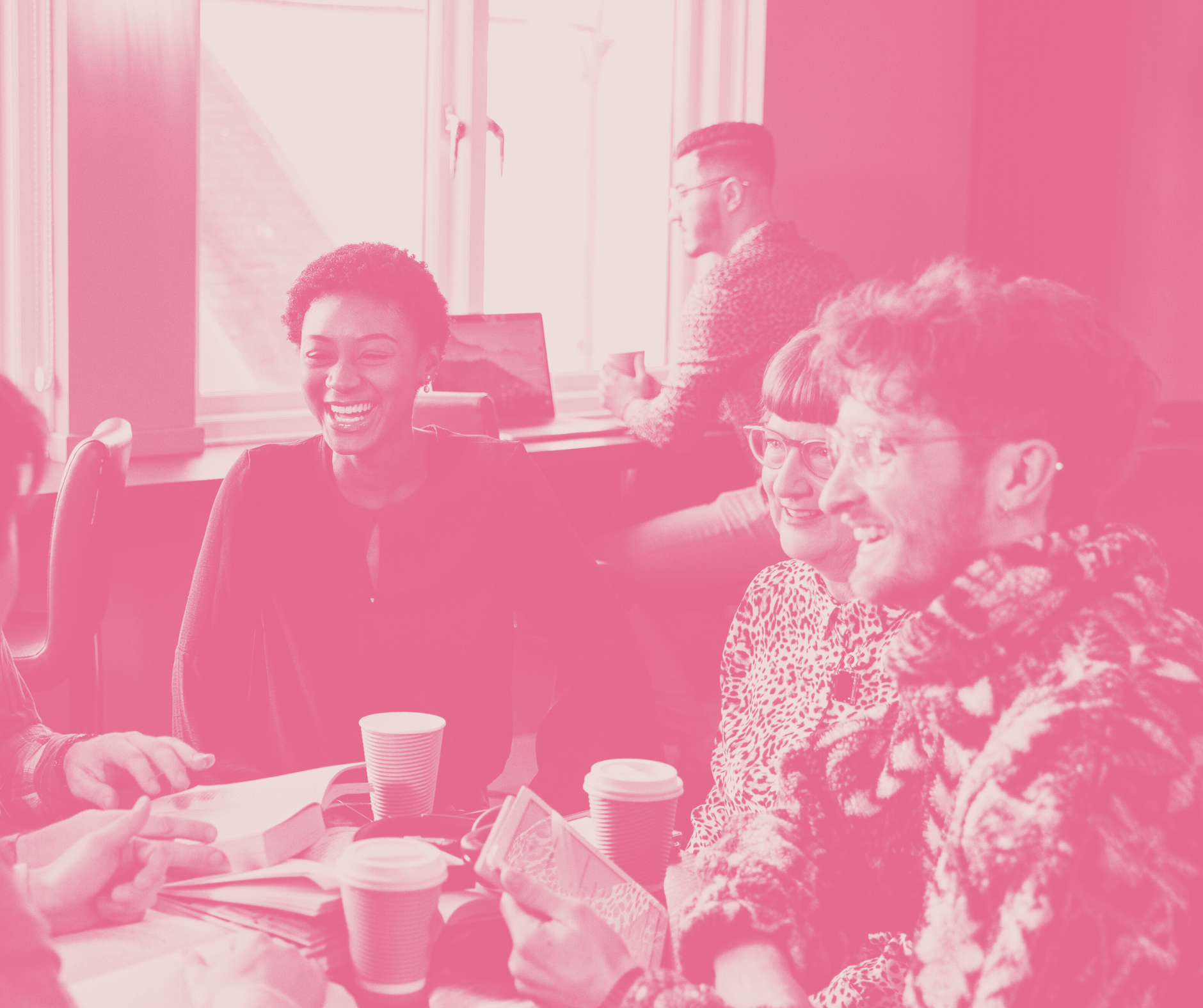 Group of diverse people laughing and talking at a meeting with coffee cups in a bright office.