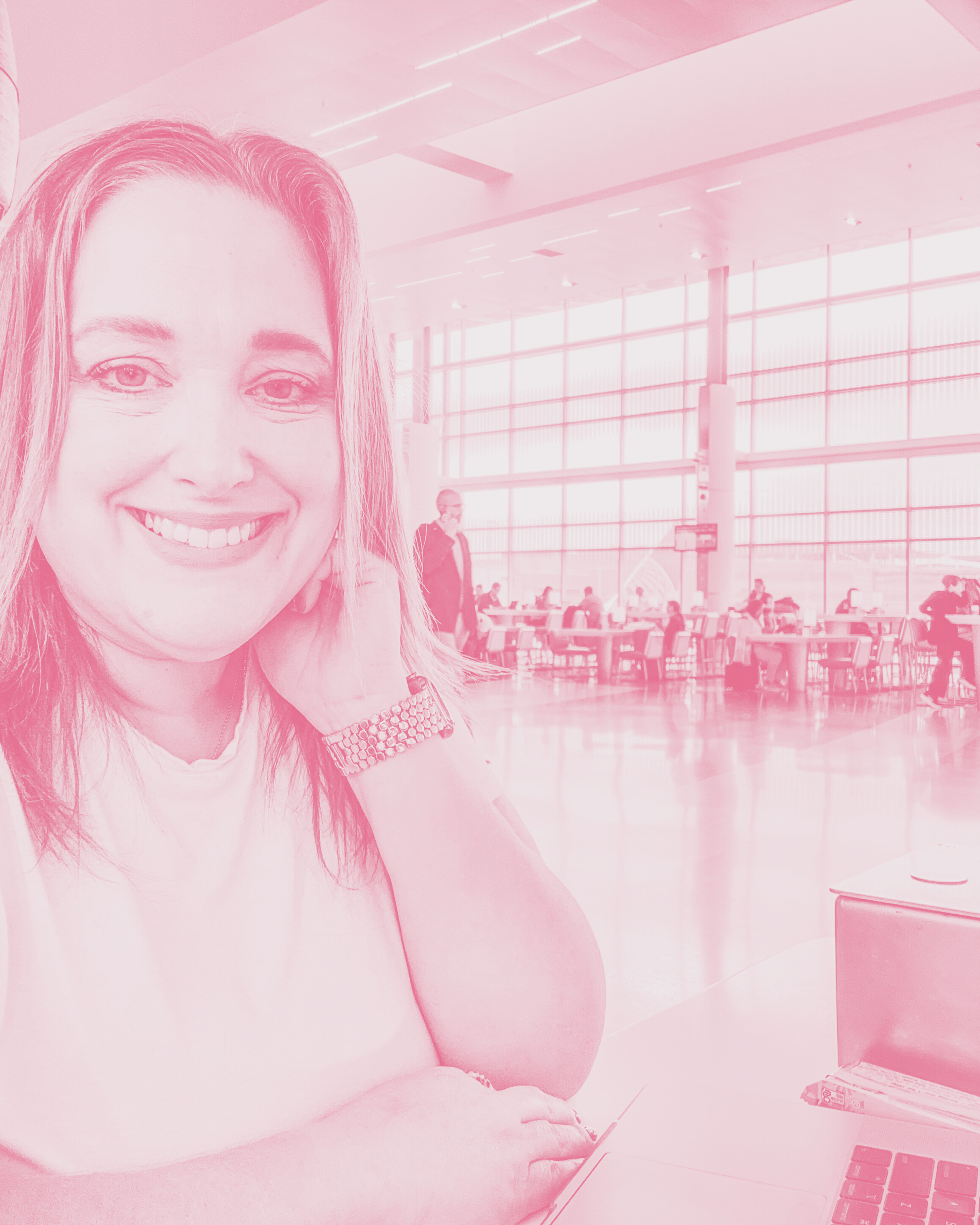 Smiling woman with shoulder-length hair sitting at a table in an airport lounge, with large floor-to-ceiling windows and people seated at tables in the background.