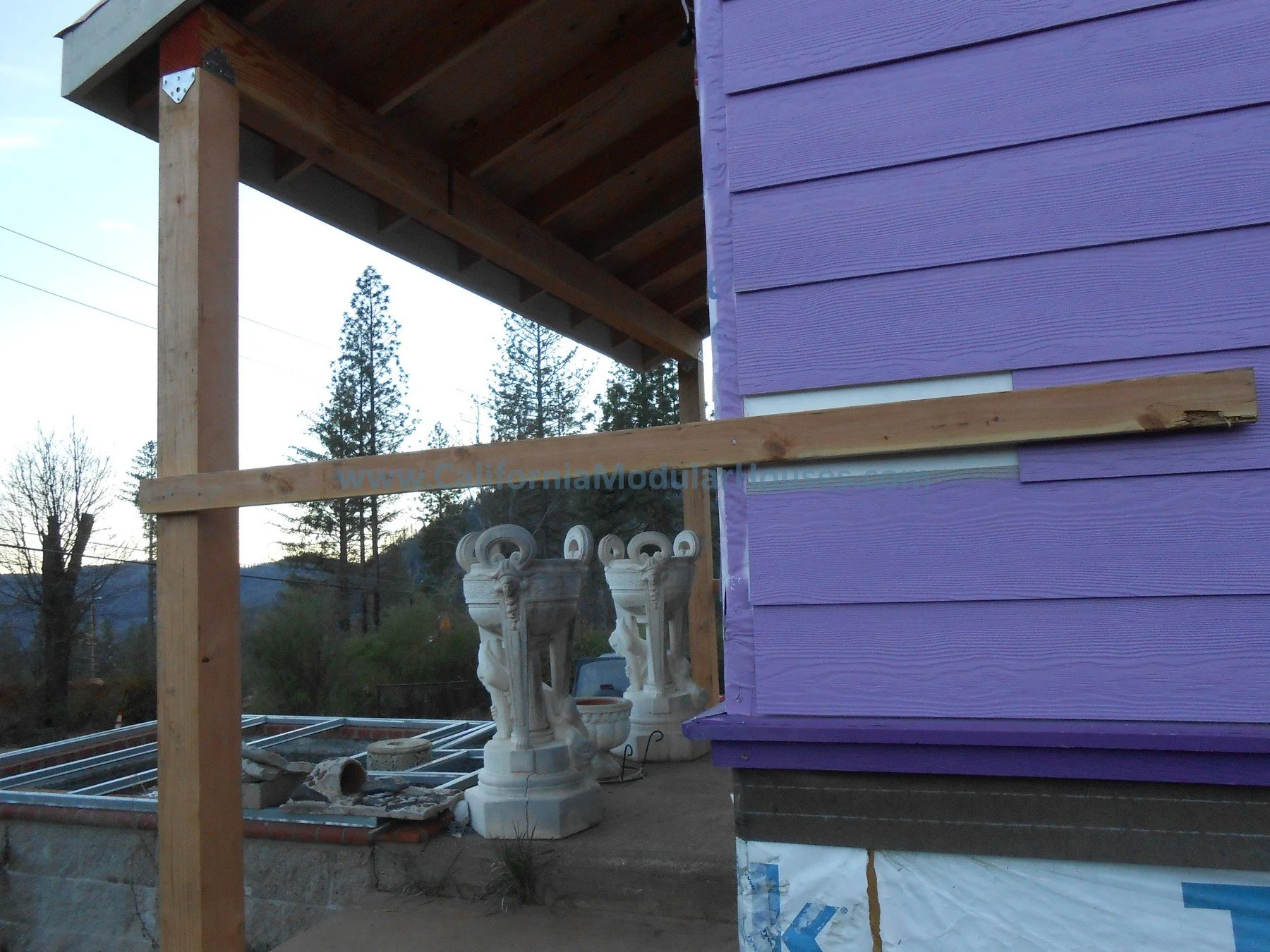 Construction site of a house with purple siding, wooden beams, construction materials, and decorative white statues of lion-like creatures.  Prefab Home California, Modular Homes in California, Modular Homes California,