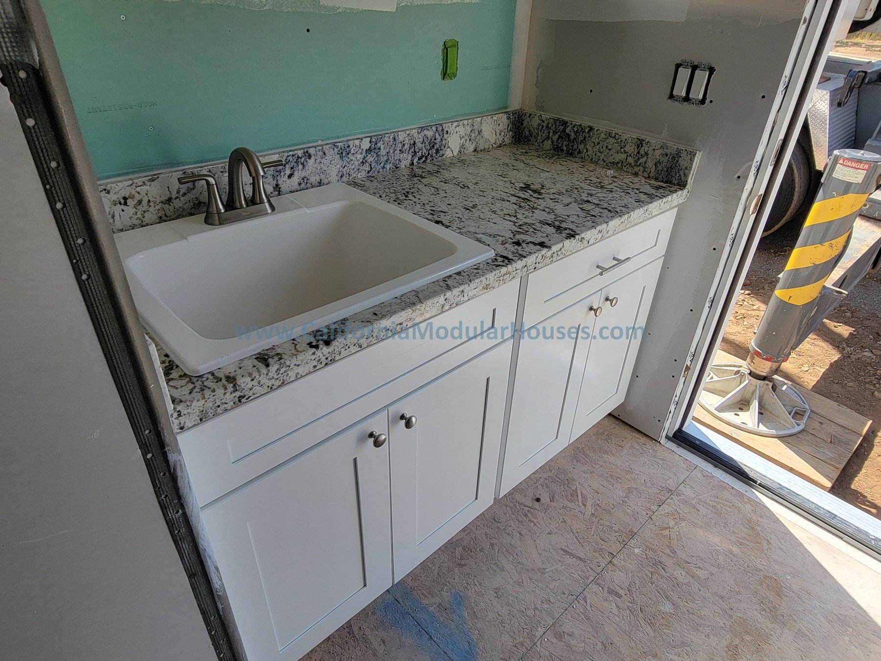 Construction site interior showing a kitchen with a granite countertop, white cabinets, a sink, and an unfinished wall with electrical outlets.