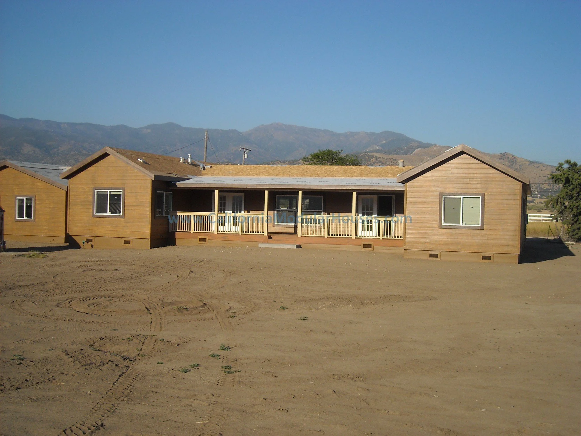 A newly constructed single-story wooden house with a central covered porch and railing, situated on a dirt lot with mountain range in the background.