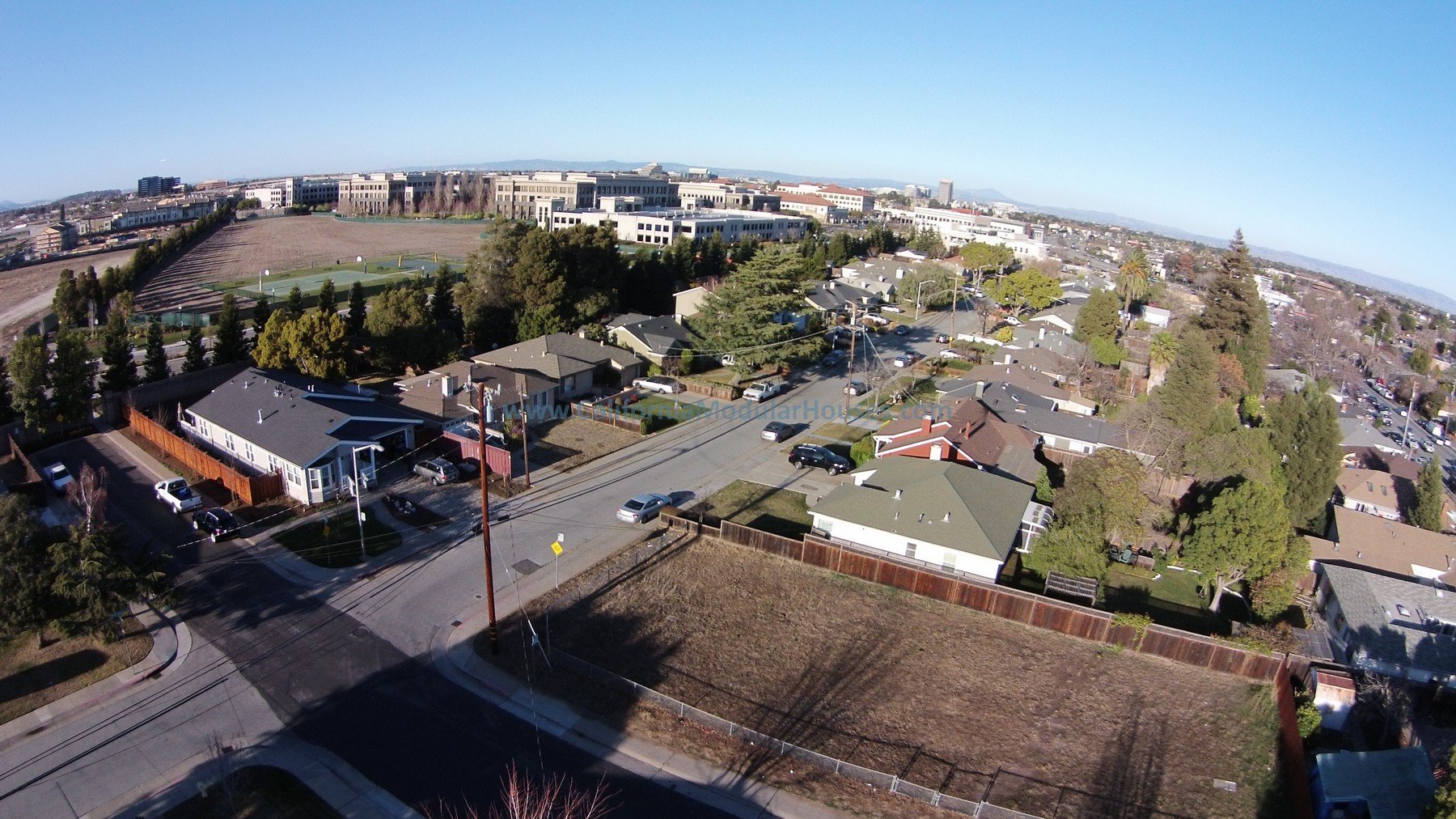 An aerial view of a neighborhood with houses, trees, and streets. In the background, there is a large parking lot and a campus-style building complex.  Modular Home CA, Modular Homes, California Modular, San Francisco Bay Area, CA.  
