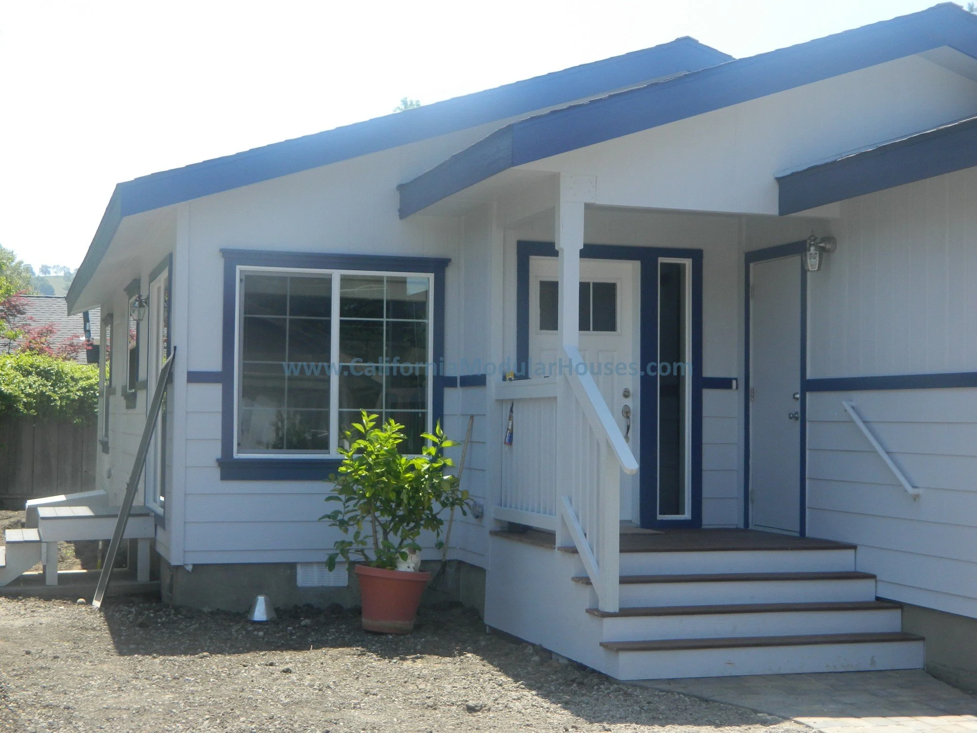 Front view of a small white house with blue trim and stairs leading to a front door, a potted plant on the porch, and a dirt yard.