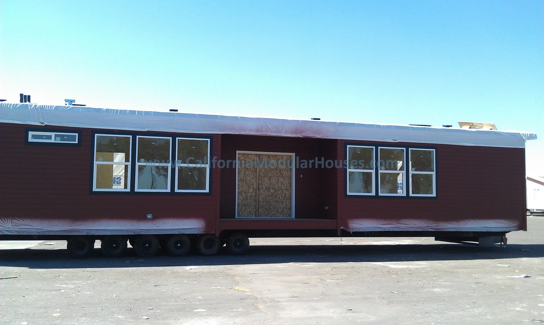 A small, red, rectangular modular house on wheels with windows and a new roof, under construction, in an open area with a clear blue sky. Modular Homes California, Modular Homes in California.