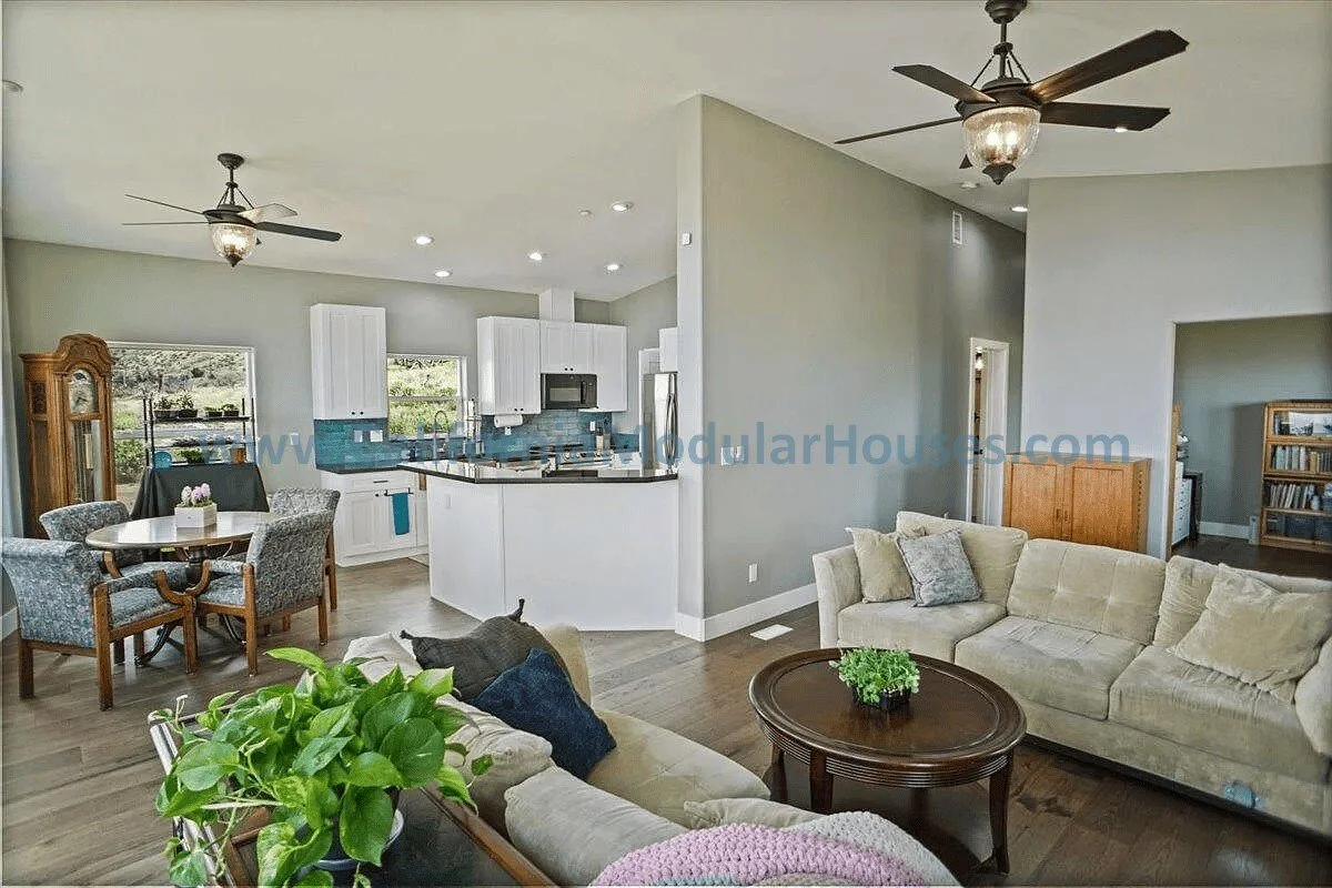 Open-concept living room and kitchen with beige sofas, a wooden dining table with four upholstered chairs, a ceiling fan, and a view of the kitchen with white cabinets, a blue tile backsplash, and stainless steel appliances.