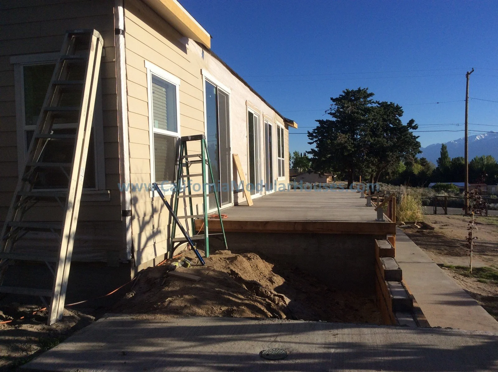 Construction site with a newly built wooden deck attached to a house, ladders and tools leaning against the house, with trees, mountains, and a clear blue sky in the background.