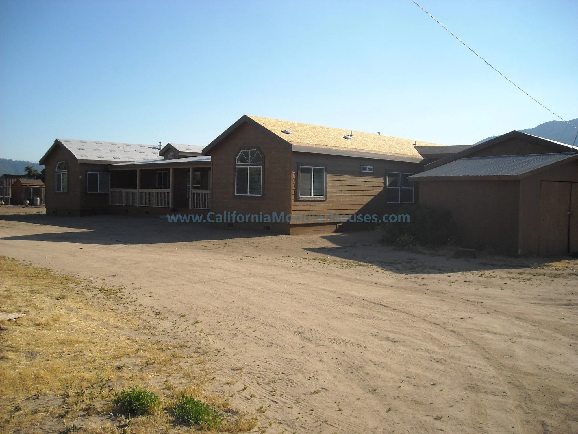 Newly constructed modular house with brown exterior, a covered porch, and multiple windows, situated on a dirt lot with a mountain in the background.  Near Lake Isabella, CA.  