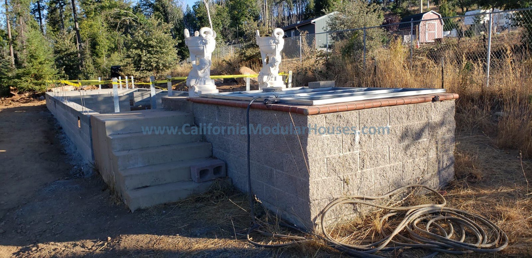 Under construction koi pond with lion statues on top, concrete stairs, cinder block foundation, and yellow caution tape in the background.  Cobb Mountain, Lake County, California.   The San Ysidro Falls model by CaliforniaModularHouses.com. 