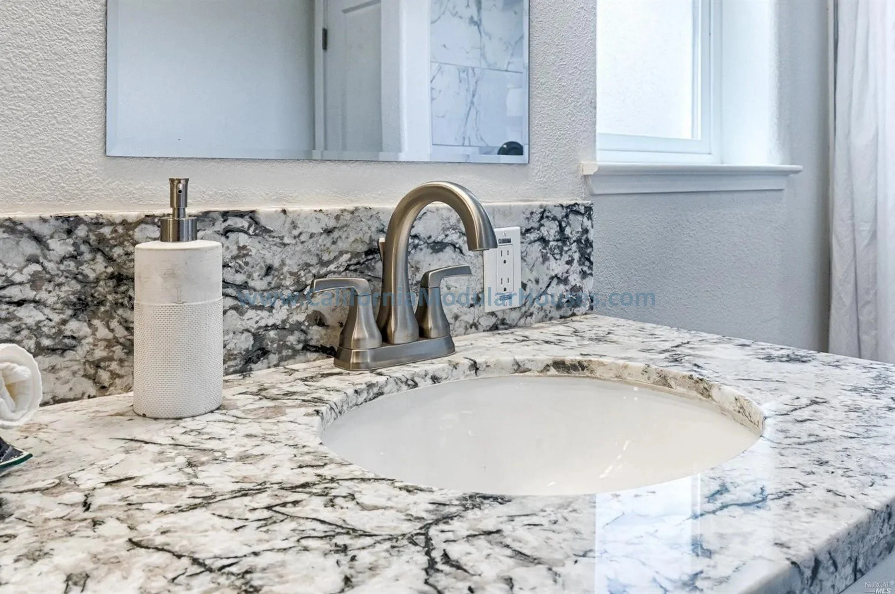 A bathroom sink with a marble countertop, a stainless steel faucet, a soap dispenser, a mirror, a window, and a wall with electrical outlets.