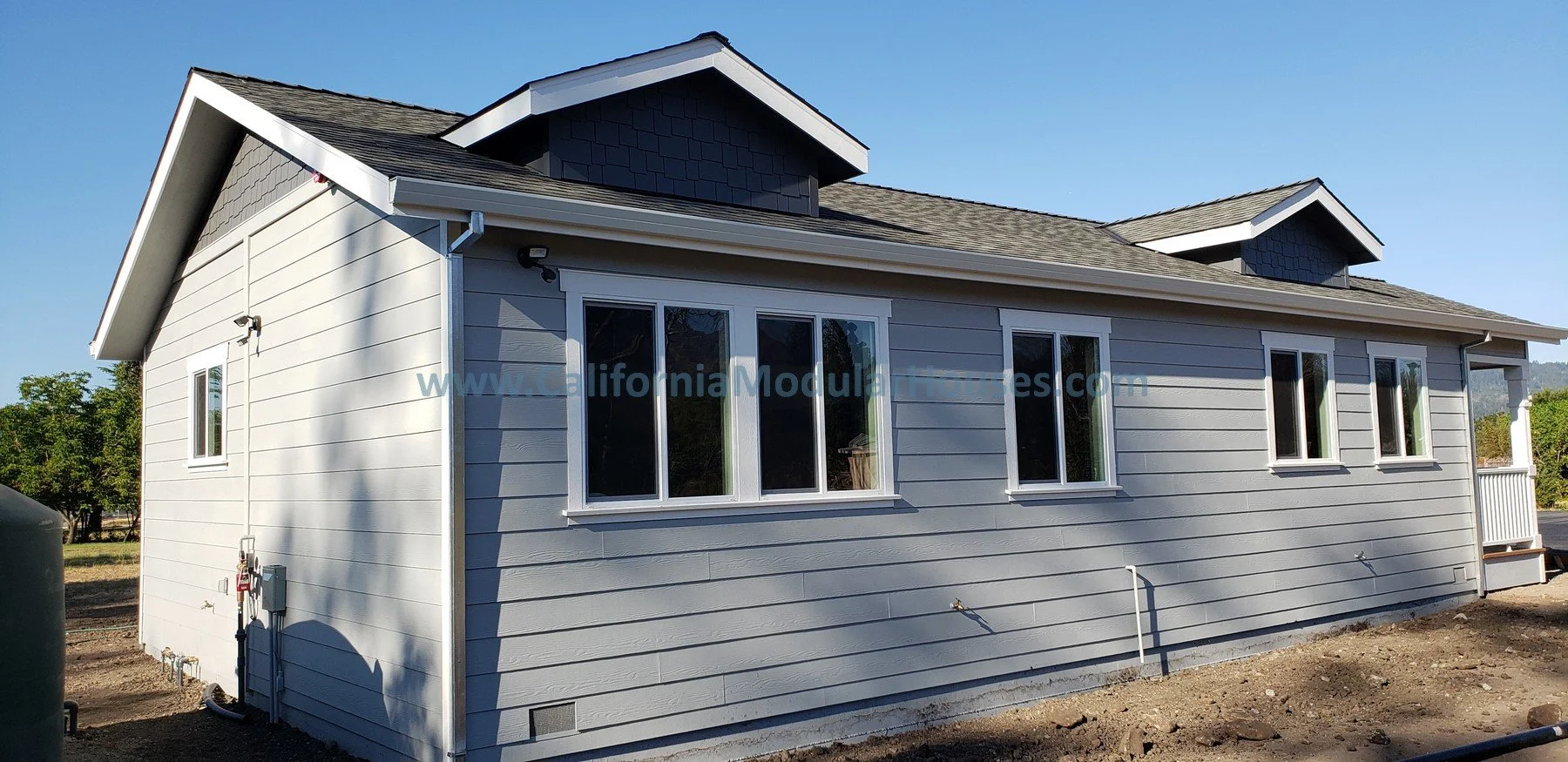Exterior of a prefab modular ADU with light gray horizontal siding, three small gable dormer windows on the roof, and multiple rectangular windows on the side facing the camera. The house is set under a clear blue sky.