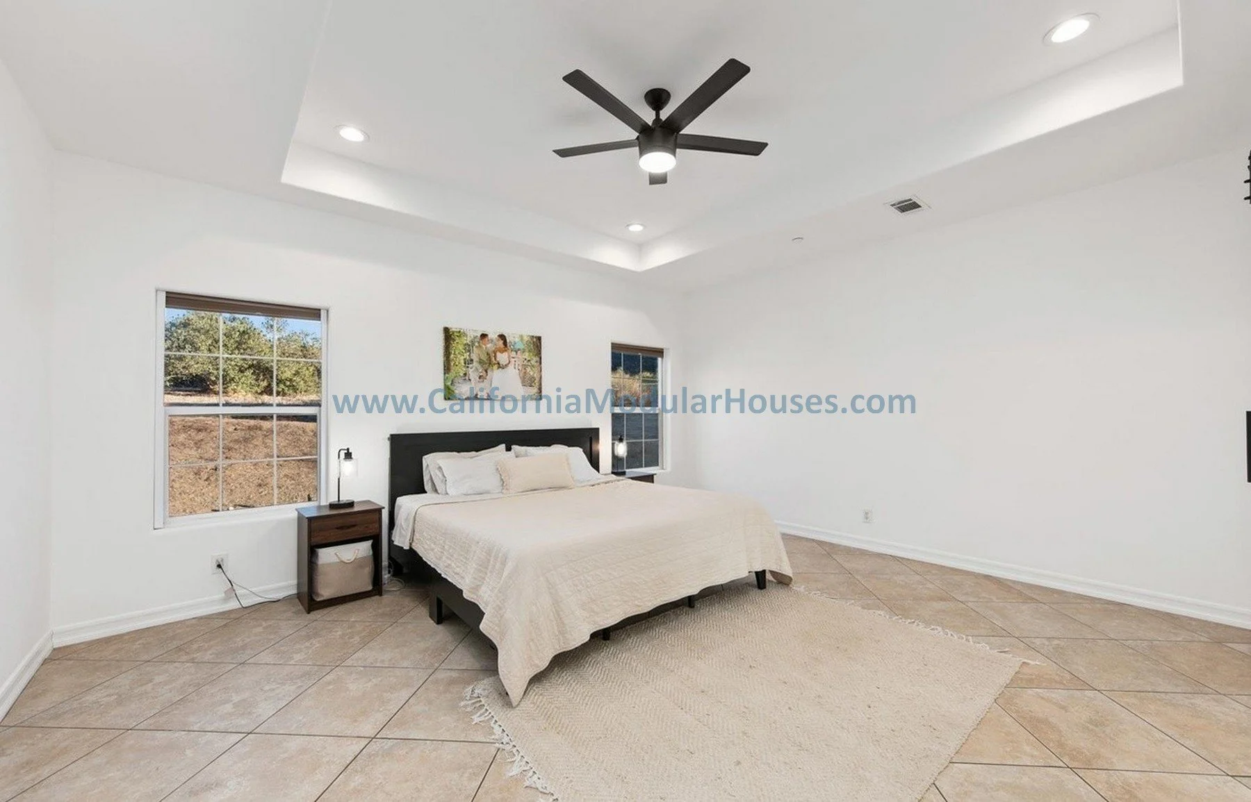 A minimalist bedroom with white walls and large tiled floor. There are two windows showing outdoor scenery, a dark wooden bed with white bedding, two small bedside tables with lamps, and a ceiling fan. A framed picture hangs above the bed.