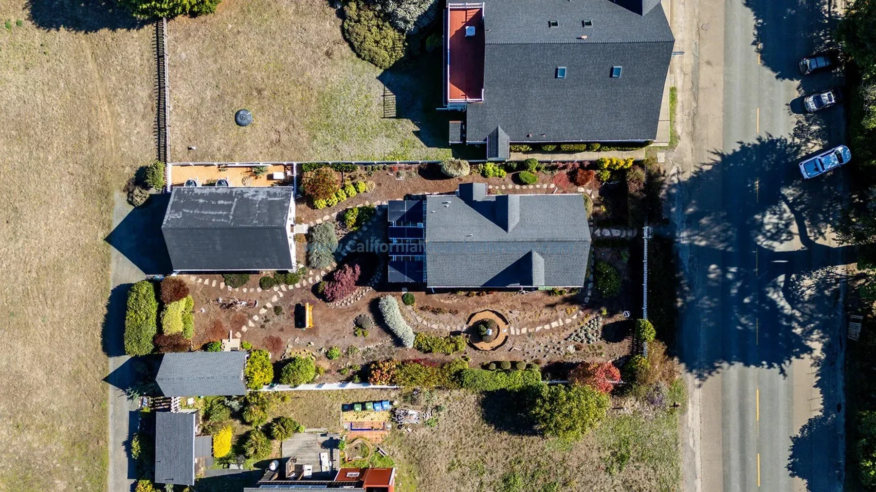 Historical. Mendocino City, CA. Prefab modular. Mendocino County, California. Aerial view of a backyard with landscaped garden, a house, and neighboring houses, with cars parked on the street and trees casting shadows.