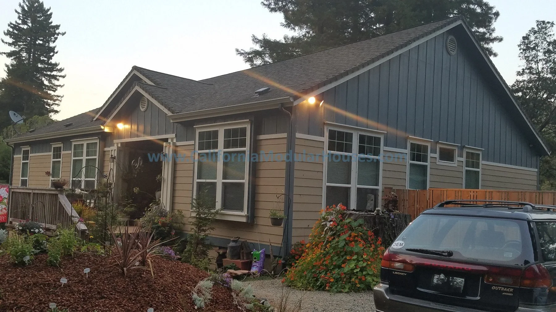 A house with blue and beige siding, multiple windows, a front garden with plants, and a black SUV parked in the driveway at dusk. Crescent City Modular Home, Modular Home on a Sloped Property. 