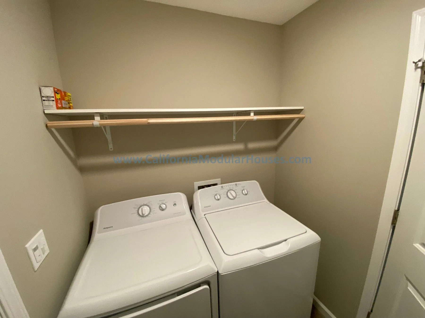 A laundry room with a white washer and dryer set, a wooden shelf above them, and a beige wall with a power outlet.  Prefab modular ADU Modular Granny Flat