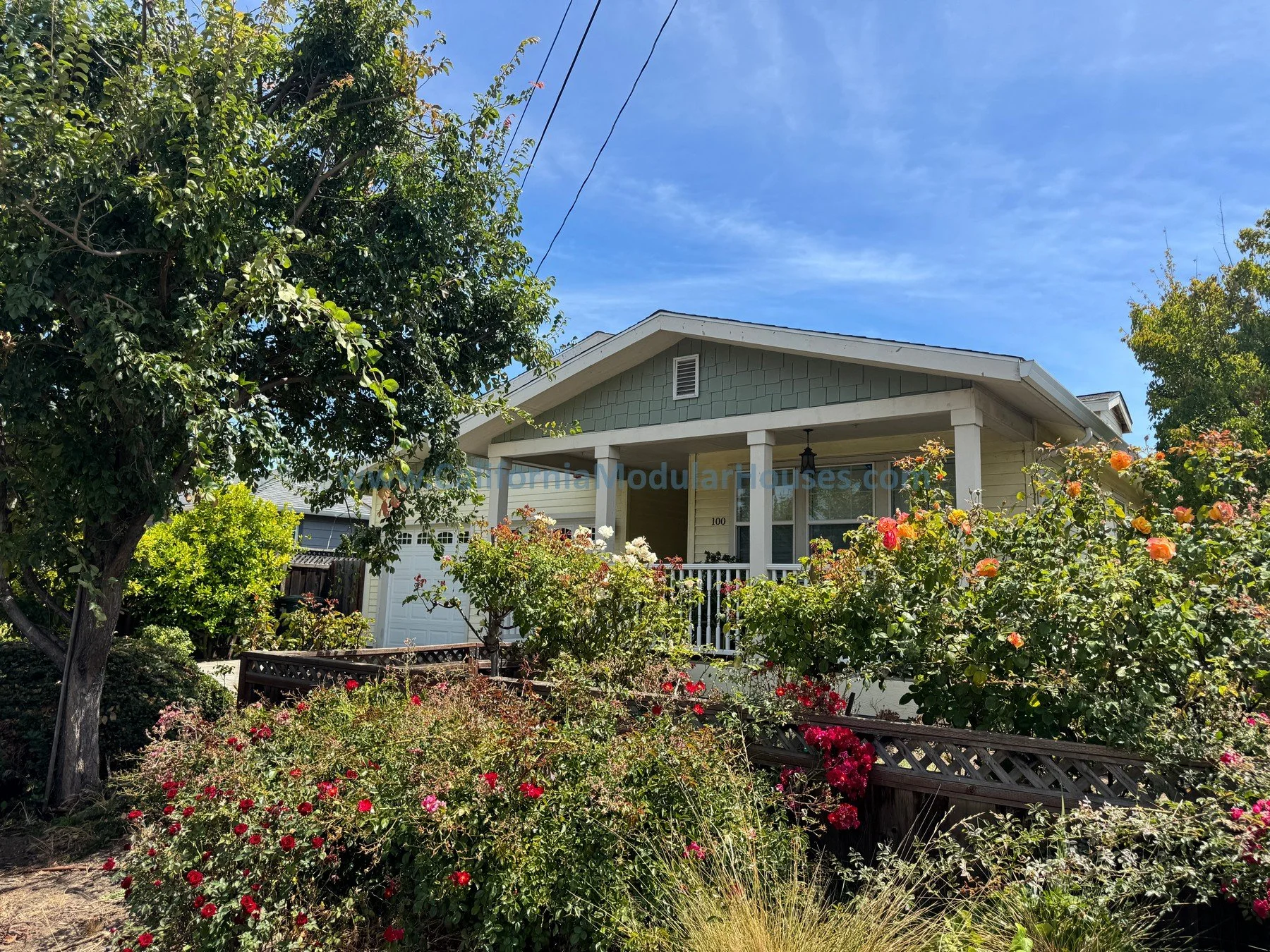 Front view of a house with a garden filled with colorful roses and lush green bushes, under a blue sky with some clouds.