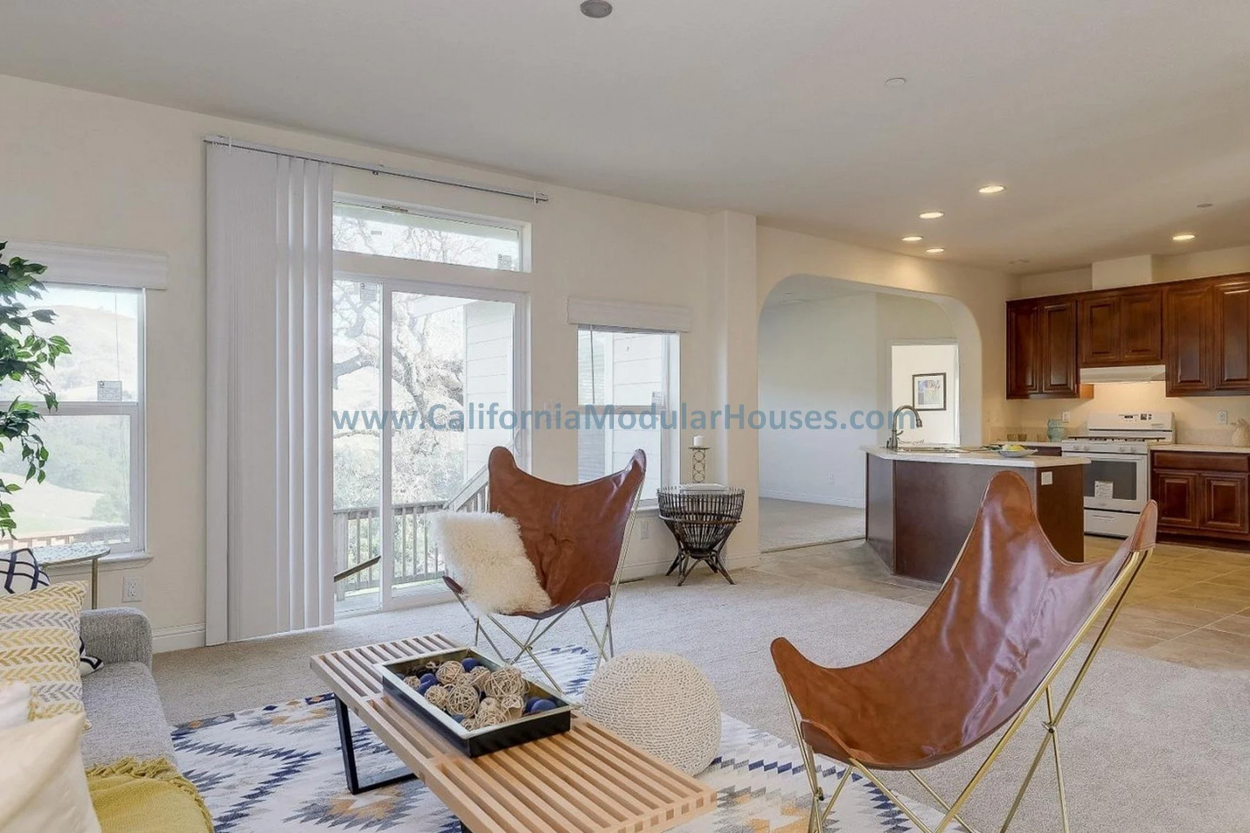 Living room with windows, leather butterfly chairs, coffee table with decorative balls, and an open kitchen with wooden cabinets and an island.  San Jose Modular Home, California Modular Homes.